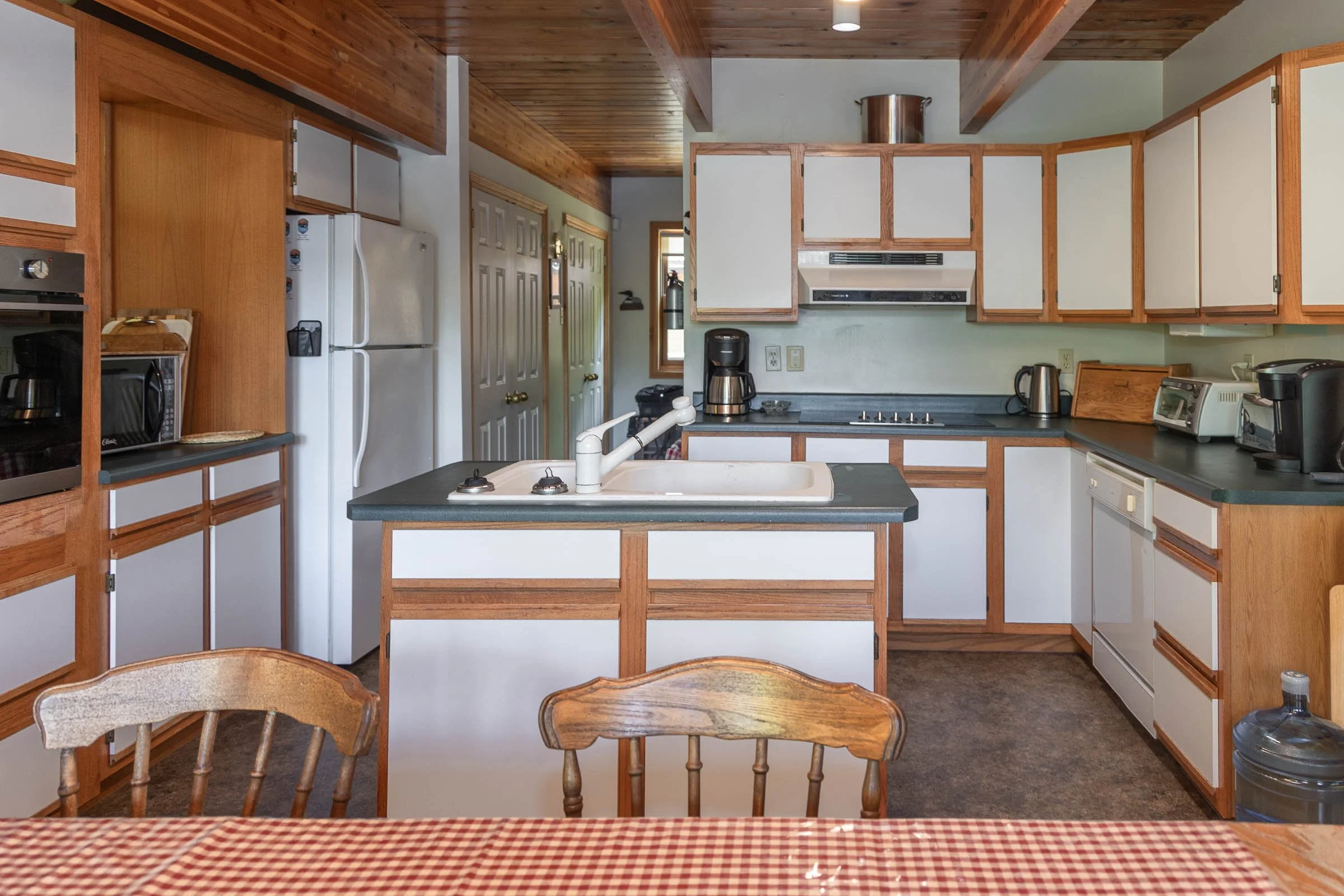 A kitchen with wooden cabinets and ceiling, white appliances including a refrigerator, oven, microwave, and toaster, and various small appliances on the countertop. There is a kitchen island with a sink, and a checkered tablecloth on a table in the f