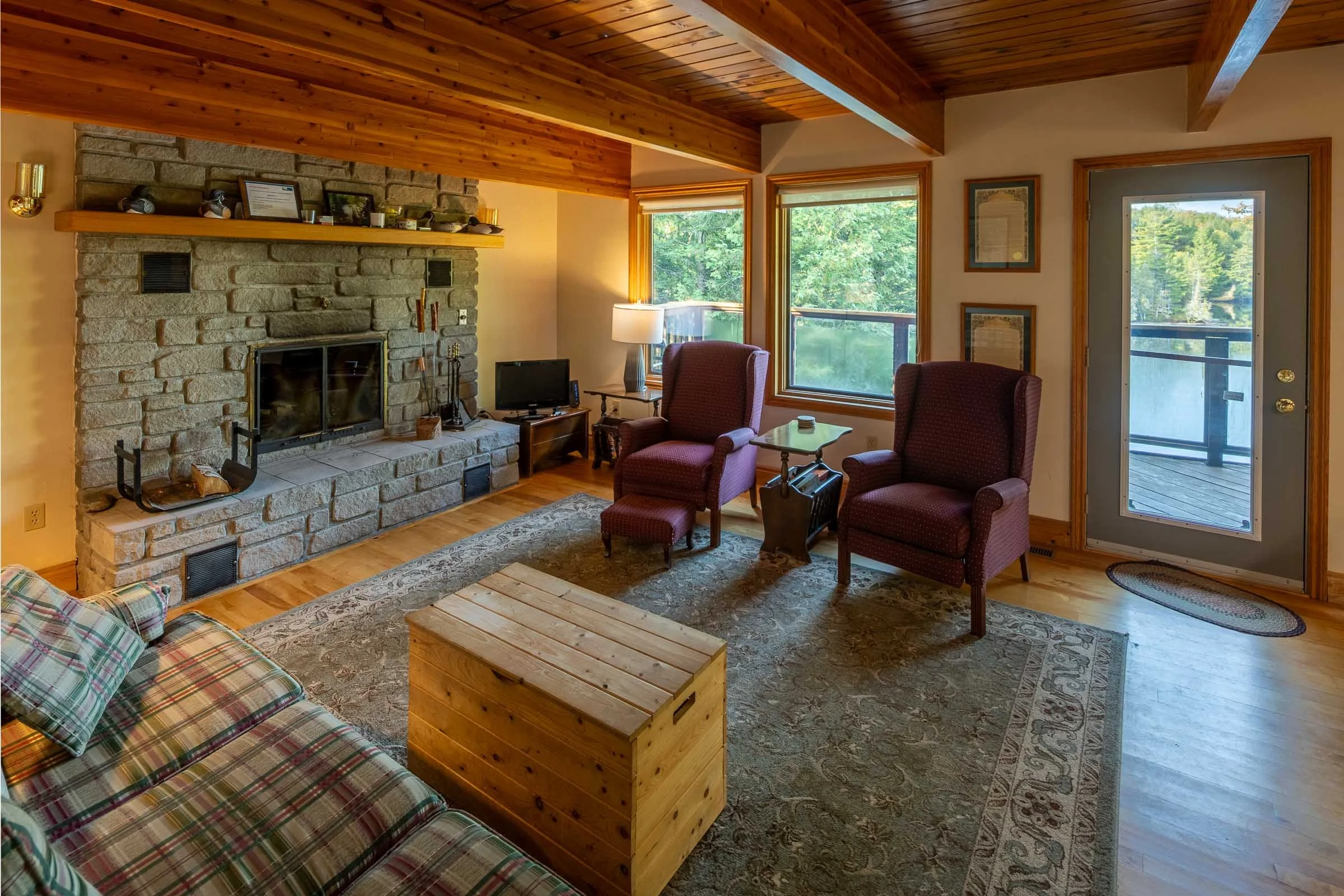 Cozy living room with wooden ceiling and large windows overlooking trees. Features a stone fireplace with a mantle, two armchairs, a small table, a TV, and a plaid sofa with a wooden coffee table.