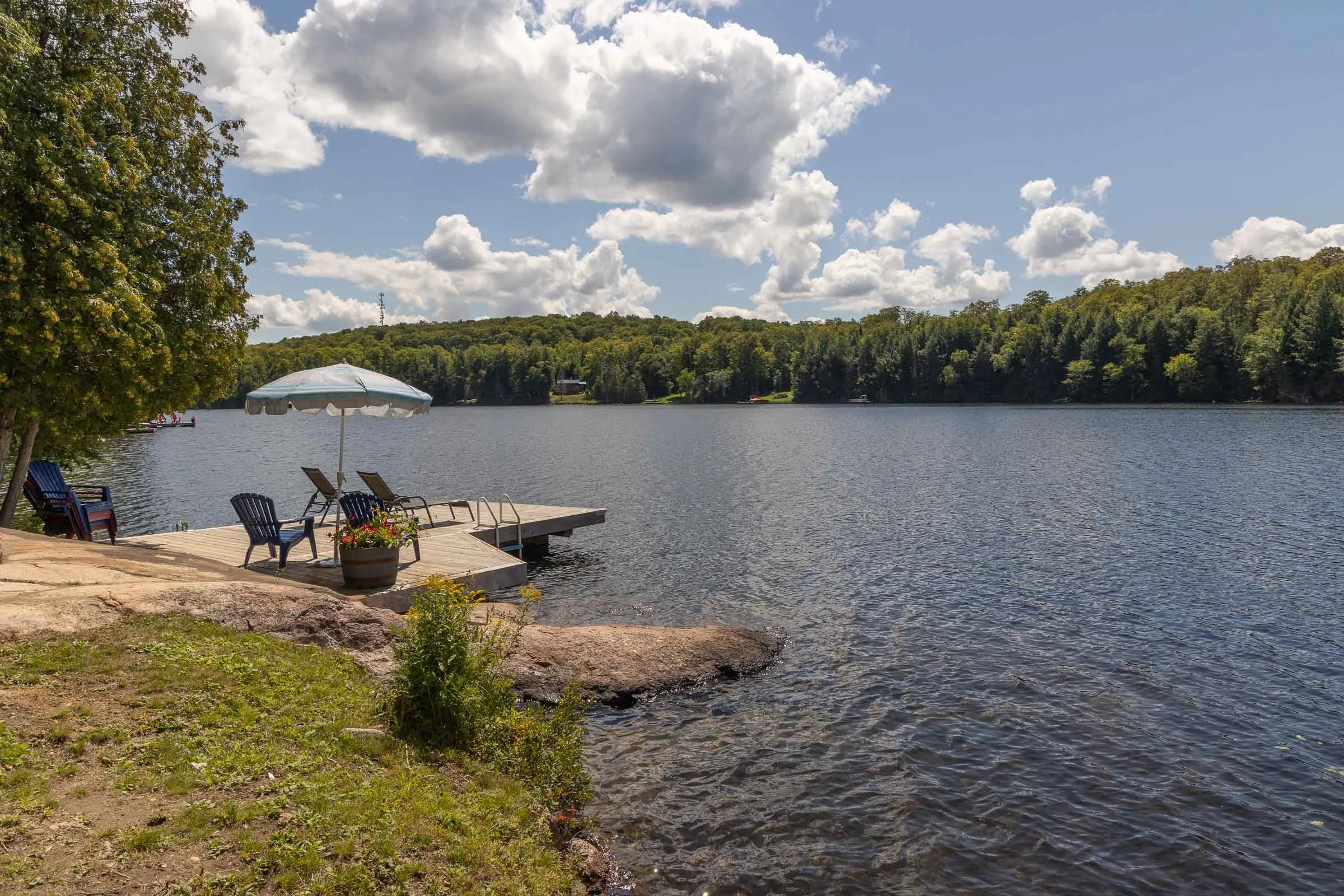A lakeside scene with a small wooden dock, beach chairs, and an umbrella. The lake is calm with reflections of the trees on the opposite shore. The sky is partly cloudy with fluffy white clouds and lush green trees cover the landscape behind the lake