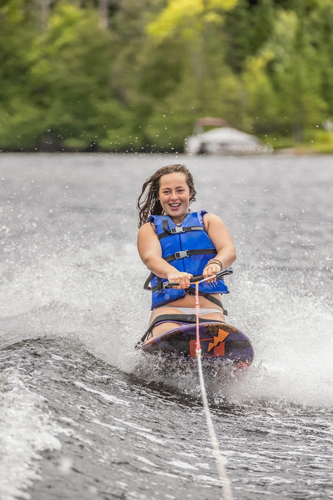 A woman with brown hair water skiing on a lake, wearing a blue life jacket, holding onto a tow rope, smiling with water splashing around her, with trees and a car in the background.