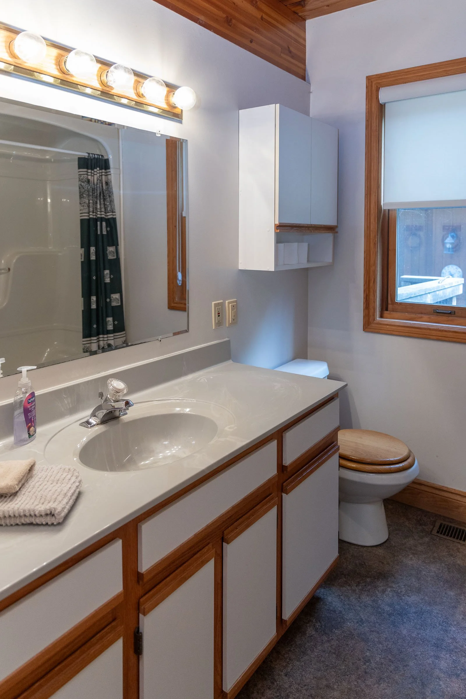 A bathroom with a white vanity and dual sinks, a large mirror with lights, a white medicine cabinet, a window with wooden trim, a toilet with a wooden lid, a gray carpet, and a shower with a striped curtain.