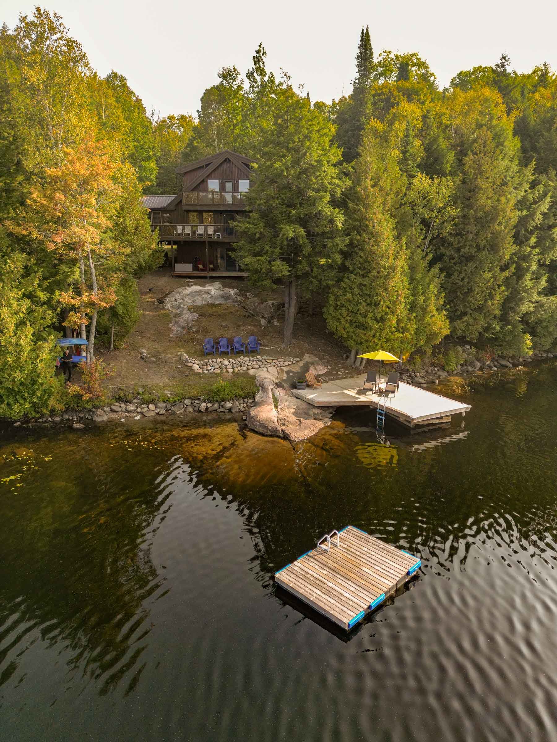 Lakeside house surrounded by trees, with a deck, a small dock in the water with a ladder, and Adirondack chairs on the shore.