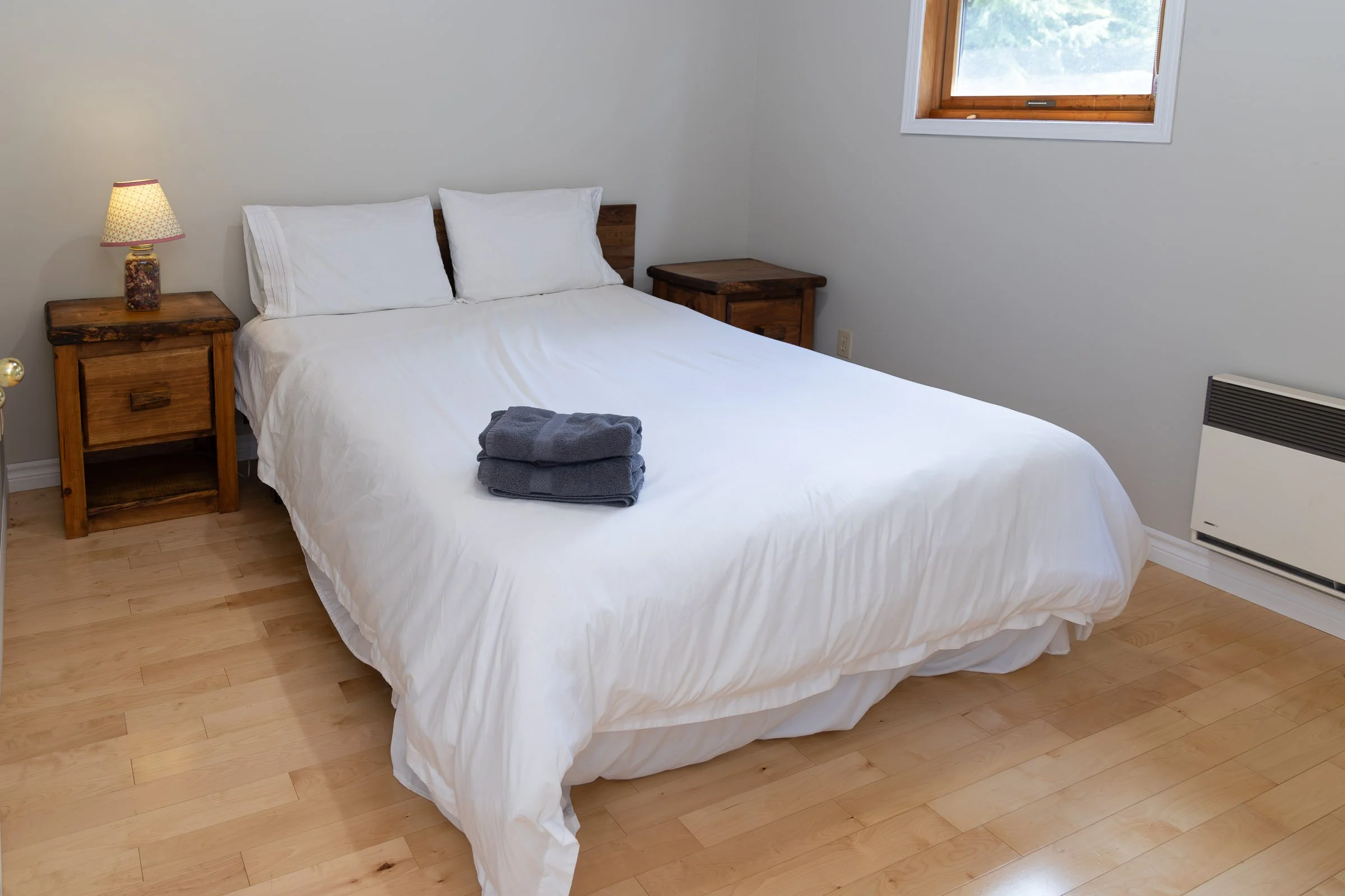 A neat bedroom with a bed covered in white sheets, two pillows, and two folded gray towels on top. There are two wooden nightstands, one with a colorful lamp on it. The room has light-colored wooden flooring, a window with a wooden frame, and a small