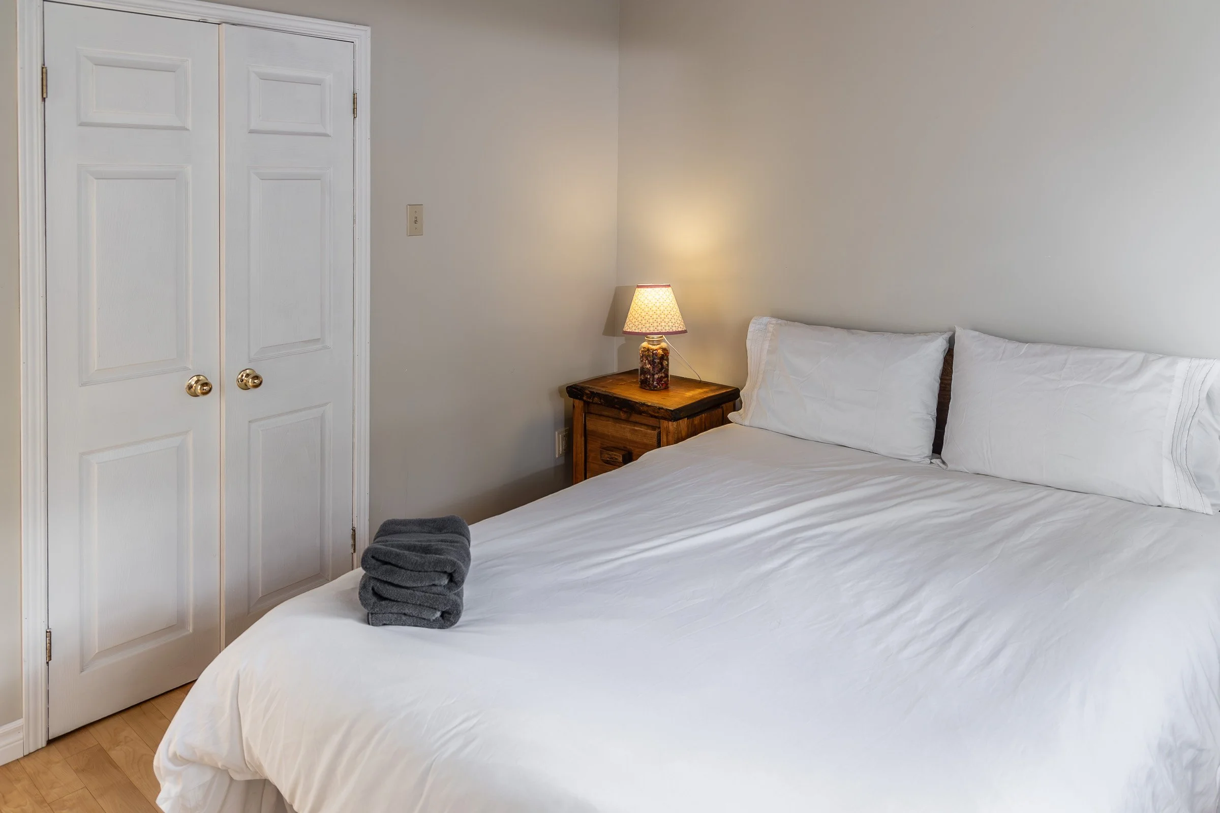 A neatly made bed with white sheets and pillows, a small wooden nightstand with a colorful lamp, and a closed closet with white doors in a simple bedroom.