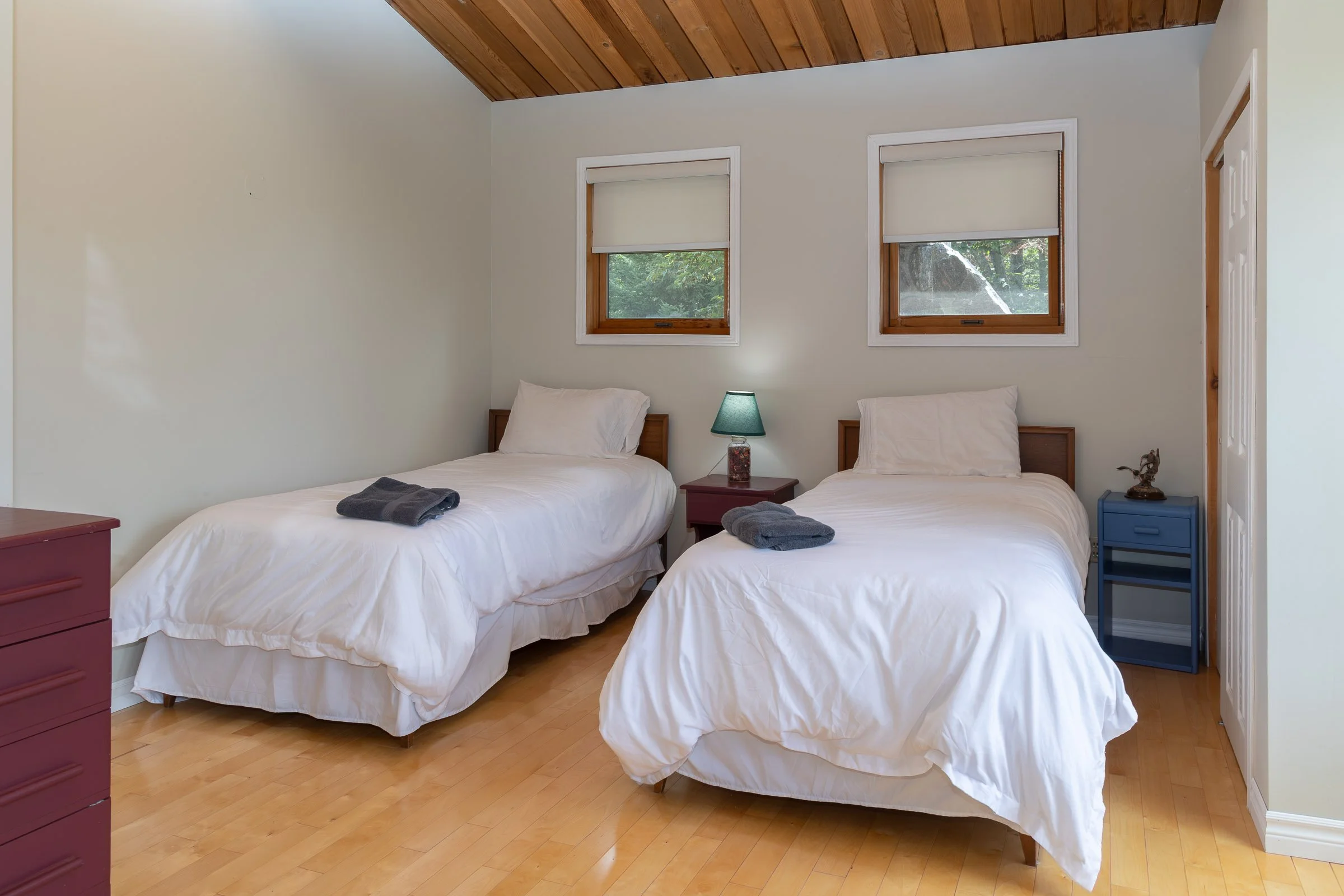 A bedroom with two twin beds covered with white bedding, each with a folded gray towel placed on top. Between the beds is a small nightstand with a lamp, and a small blue shelf or side table on the right side. The room has light-colored walls, two wi