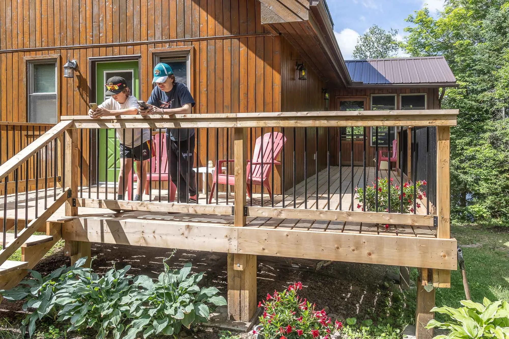 Two boys standing on a wooden deck outside a house, looking at their phones. The house has a wooden exterior with a green door and multiple windows. There is outdoor furniture including pink chairs on the deck, and greenery with trees and plants surr