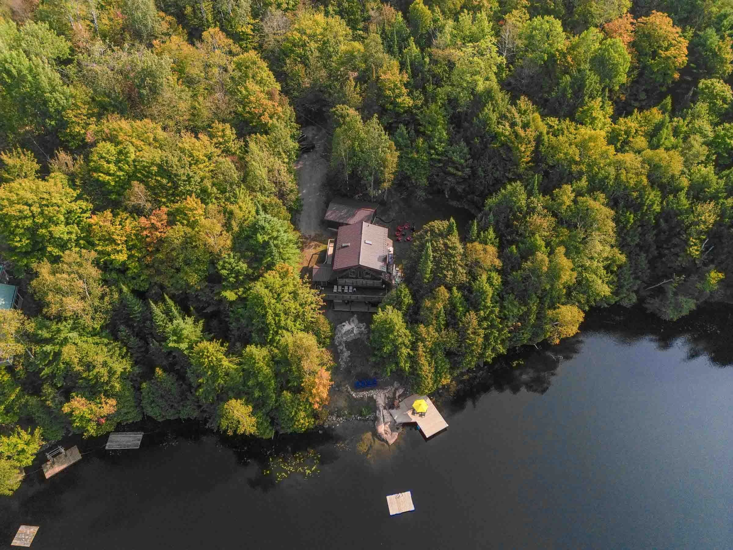 An aerial view of a lake surrounded by a dense forest with trees showing green, yellow, and orange foliage. There are a few houses with docks along the shoreline, and the water is calm.