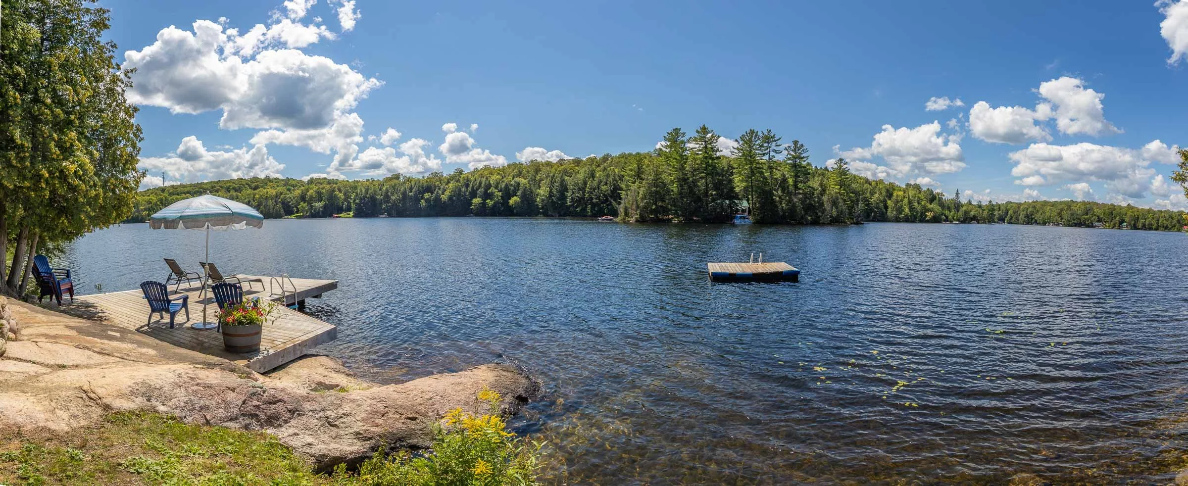 A lakeside view with a small wooden dock, seating area with chairs and an umbrella, a tree on the left, water with a floating dock, and a forested shoreline under a partly cloudy blue sky.