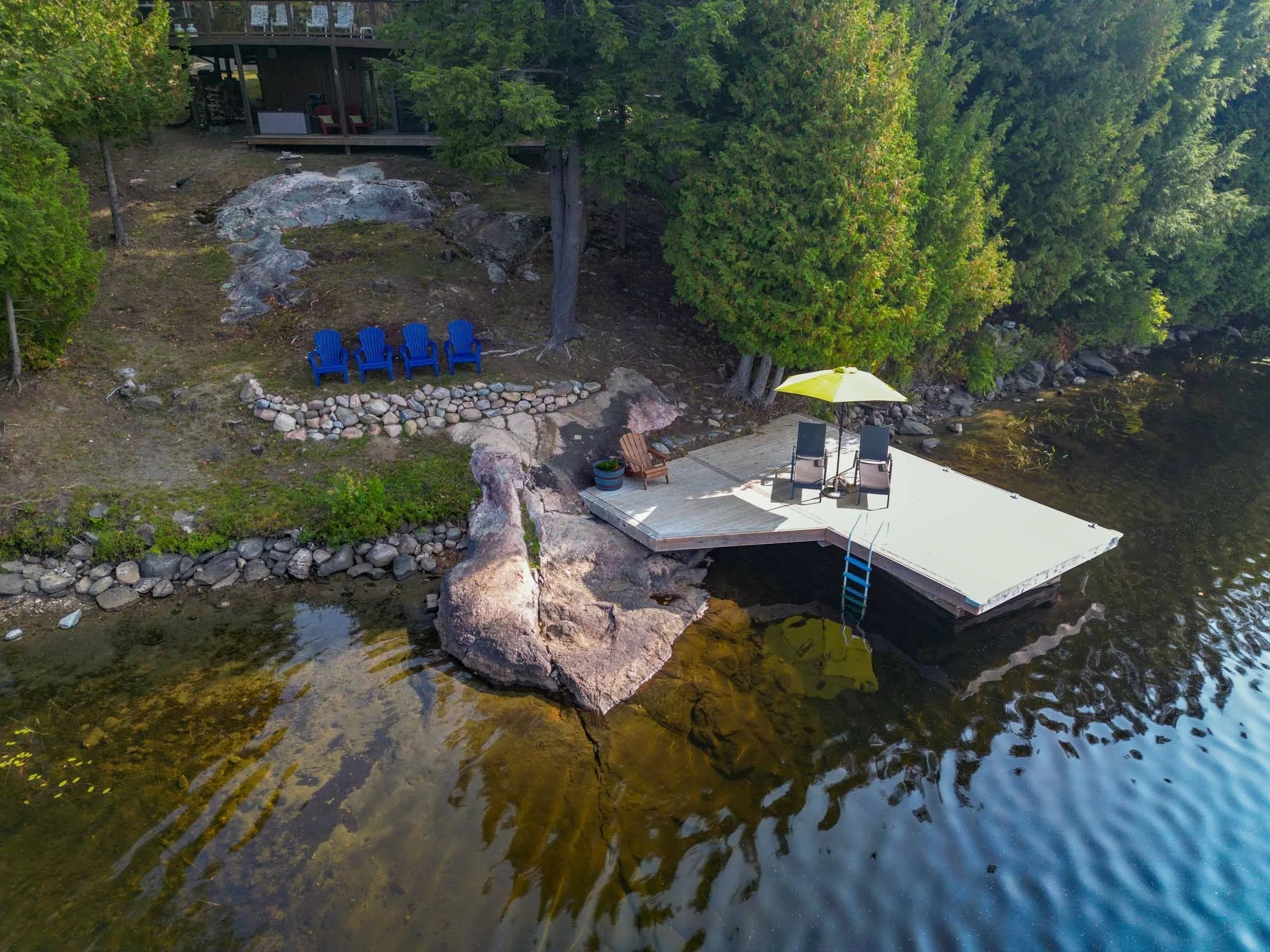 Overhead view of a lakeside patio with four blue Adirondack chairs, a small rock arrangement, a yellow umbrella, a pool, and a wooded area with large rocks and trees.