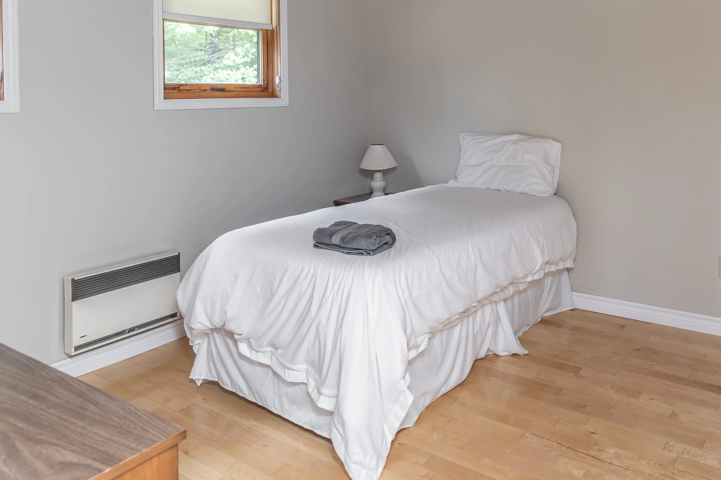 A simple bedroom with a white bed covered in a white bedspread and matching pillow. A folded gray towel rests on the bed. There is a small white nightstand with a white lamp. Two windows with wooden frames are on the wall, showing green trees outside