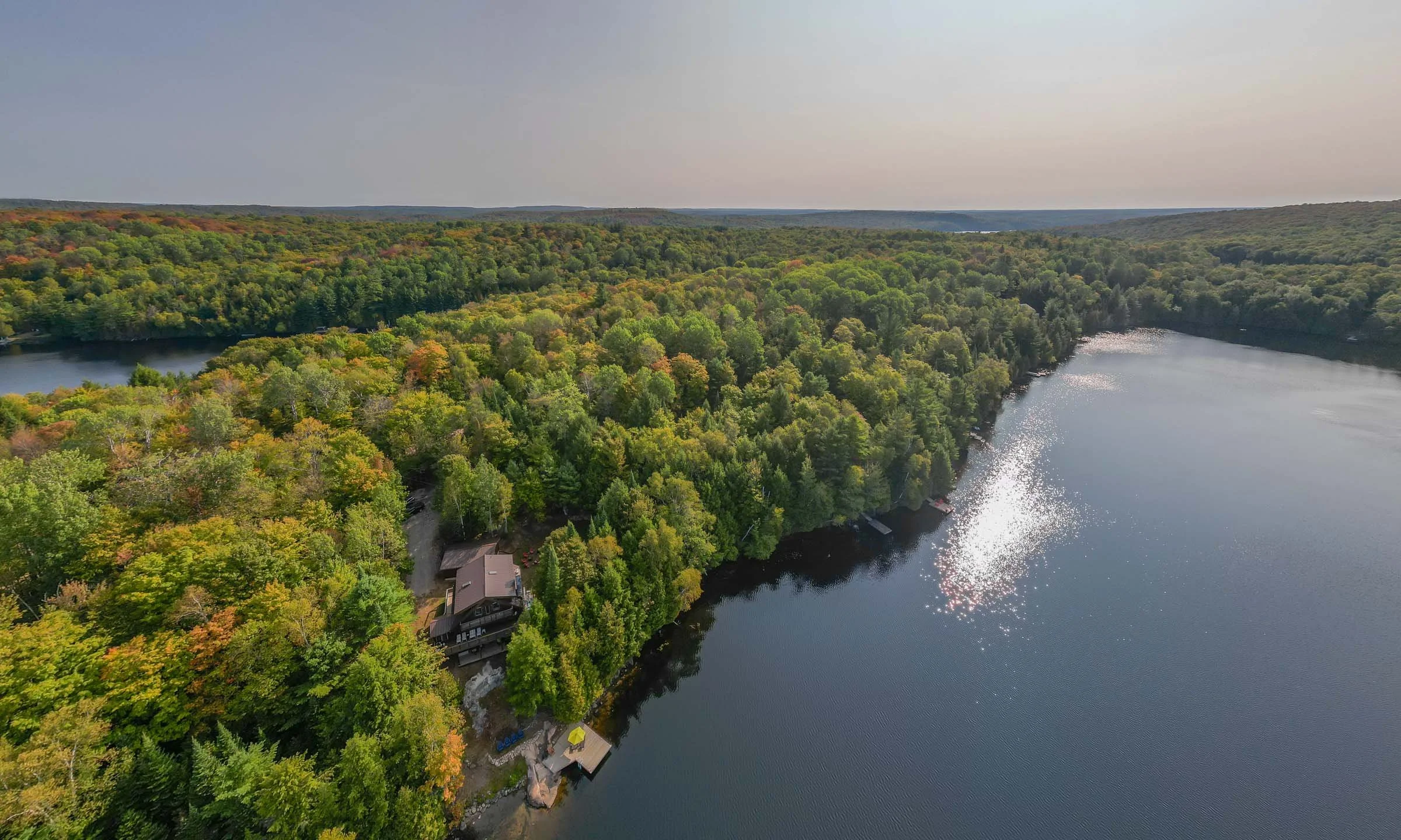 Aerial view of a large lake surrounded by densely forested hills with trees showing early signs of autumn colors. There are buildings and docks near the lake's edge, and the sky above is clear with a slight haze.