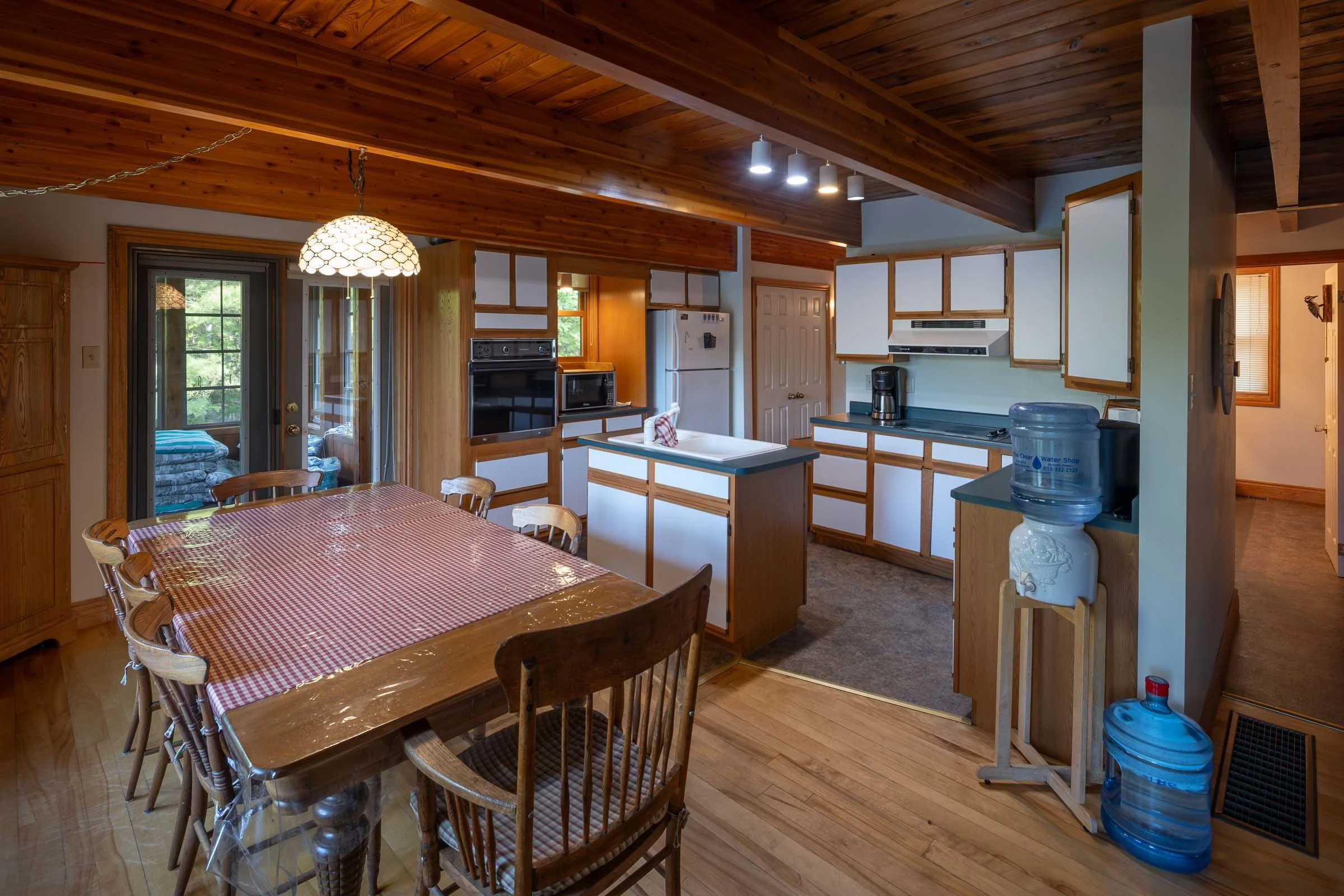 Wood-paneled kitchen and dining area with a checkered tablecloth on a wooden table, surrounded by wooden chairs, and a water cooler in the corner. Kitchen cabinets with white panels and a black stovetop are visible, with a door leading outside and natural light coming through the windows.