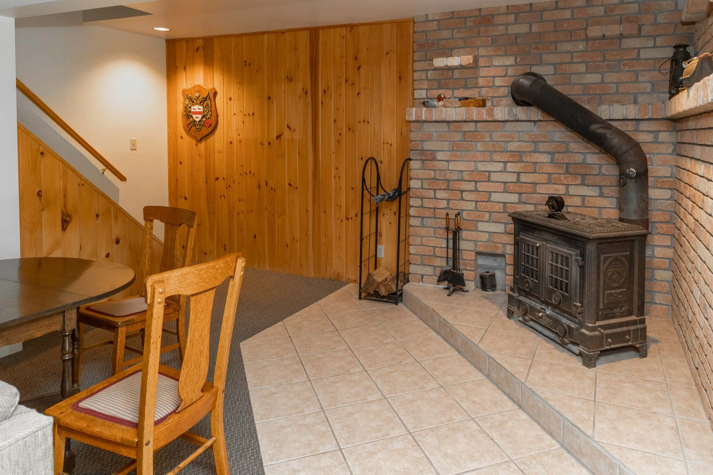 Living room with wooden panel wall, brick fireplace with wood stove, black pipe, and fireplace tools. Wooden chairs around a round table. Coat rack next to fireplace. Coat of arms with eagle and shield hanging on wall. Carpeted area and tile floor.