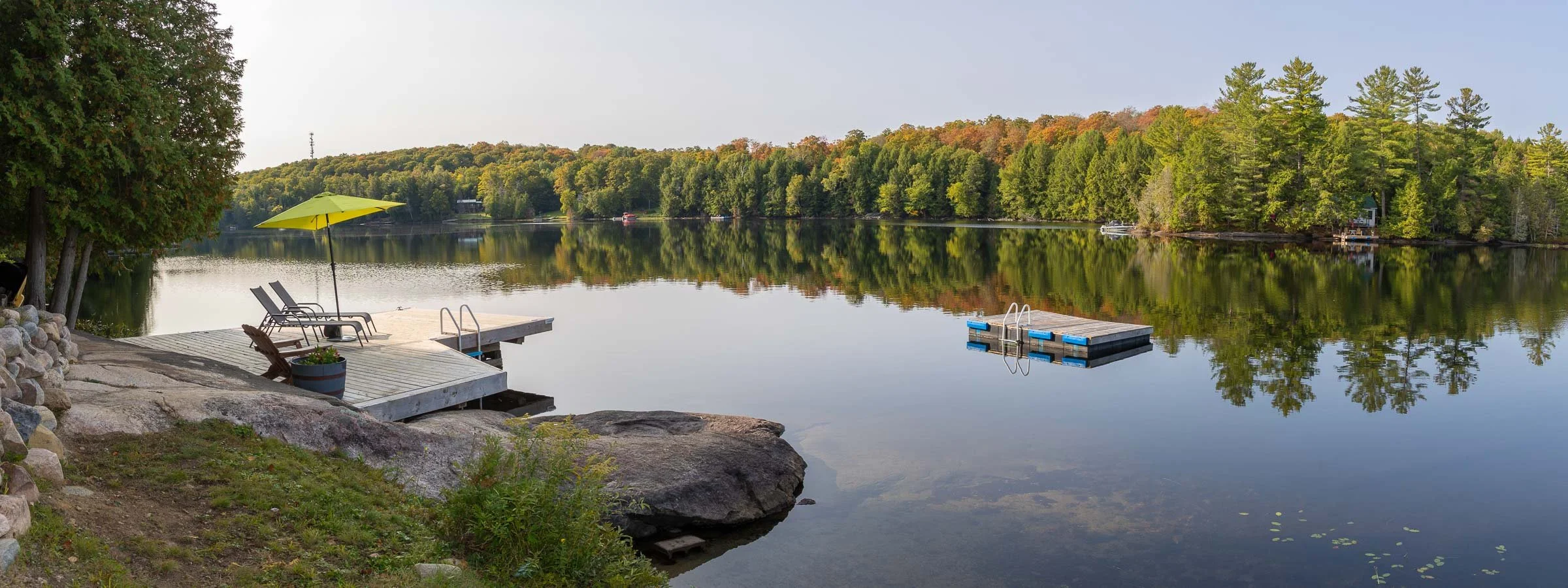 A lakeside scene with a wooden dock, two lounge chairs, a yellow umbrella, and a floating dock surrounded by trees with fall foliage.