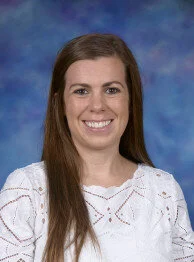 Professional portrait of a woman with long brown hair, smiling, wearing a white blouse with a textured pattern, against a blue patterned background.