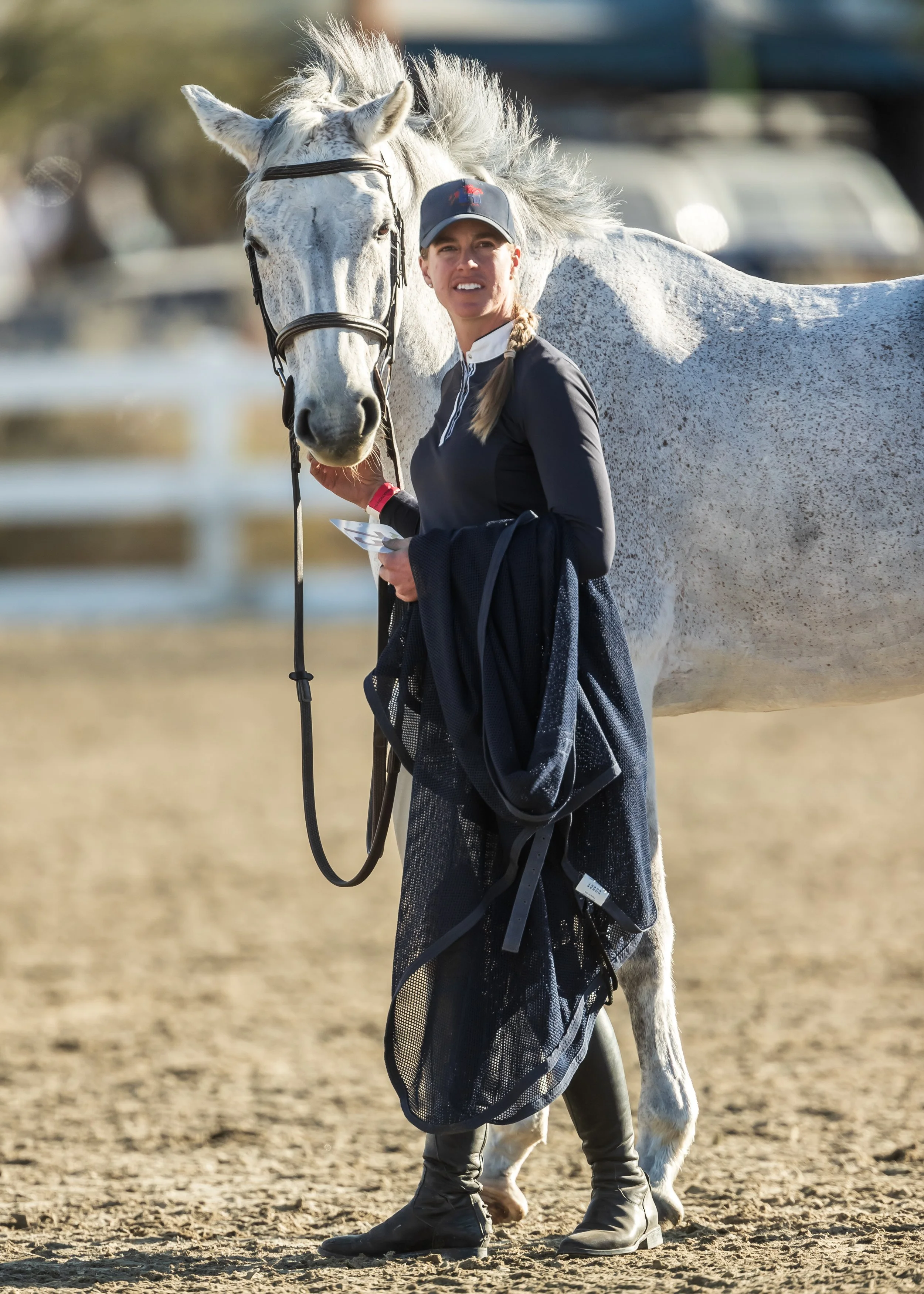 Woman standing next to a white horse outdoors on a dirt field, holding the horse's reins.