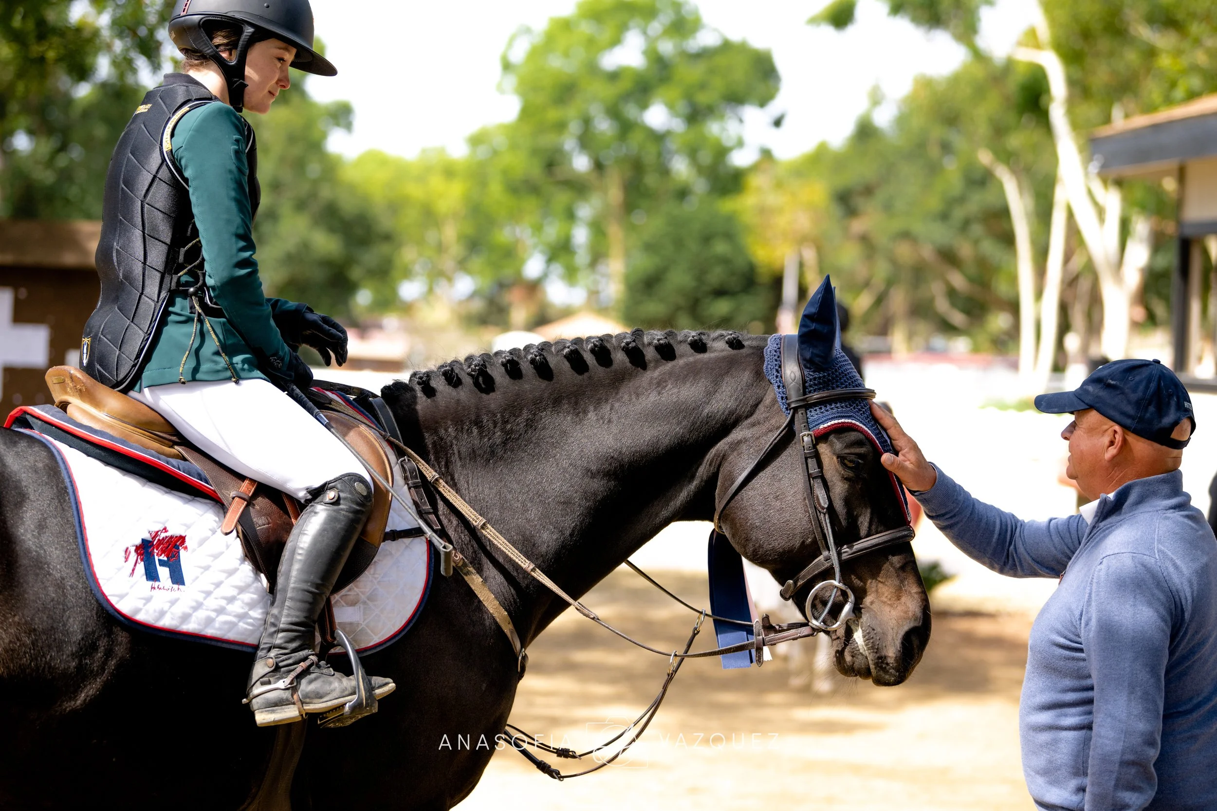 A woman with a helmet and riding gear is sitting on a horse, and a man is holding the horse's bridle, helping her prepare for riding outdoors in a wooded area.