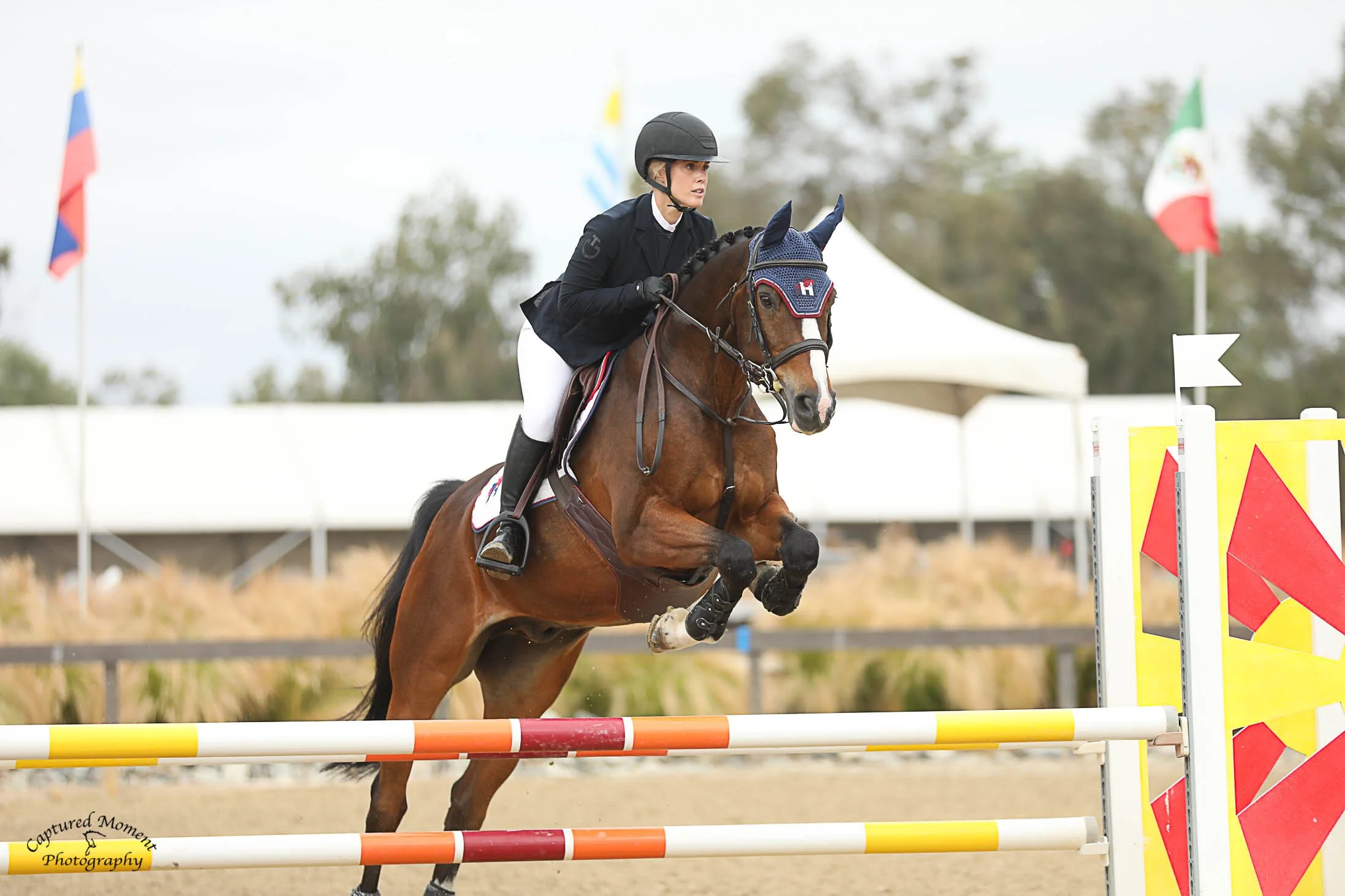 A female equestrian riding a horse and jumping over an obstacle at an outdoor show jumping competition.