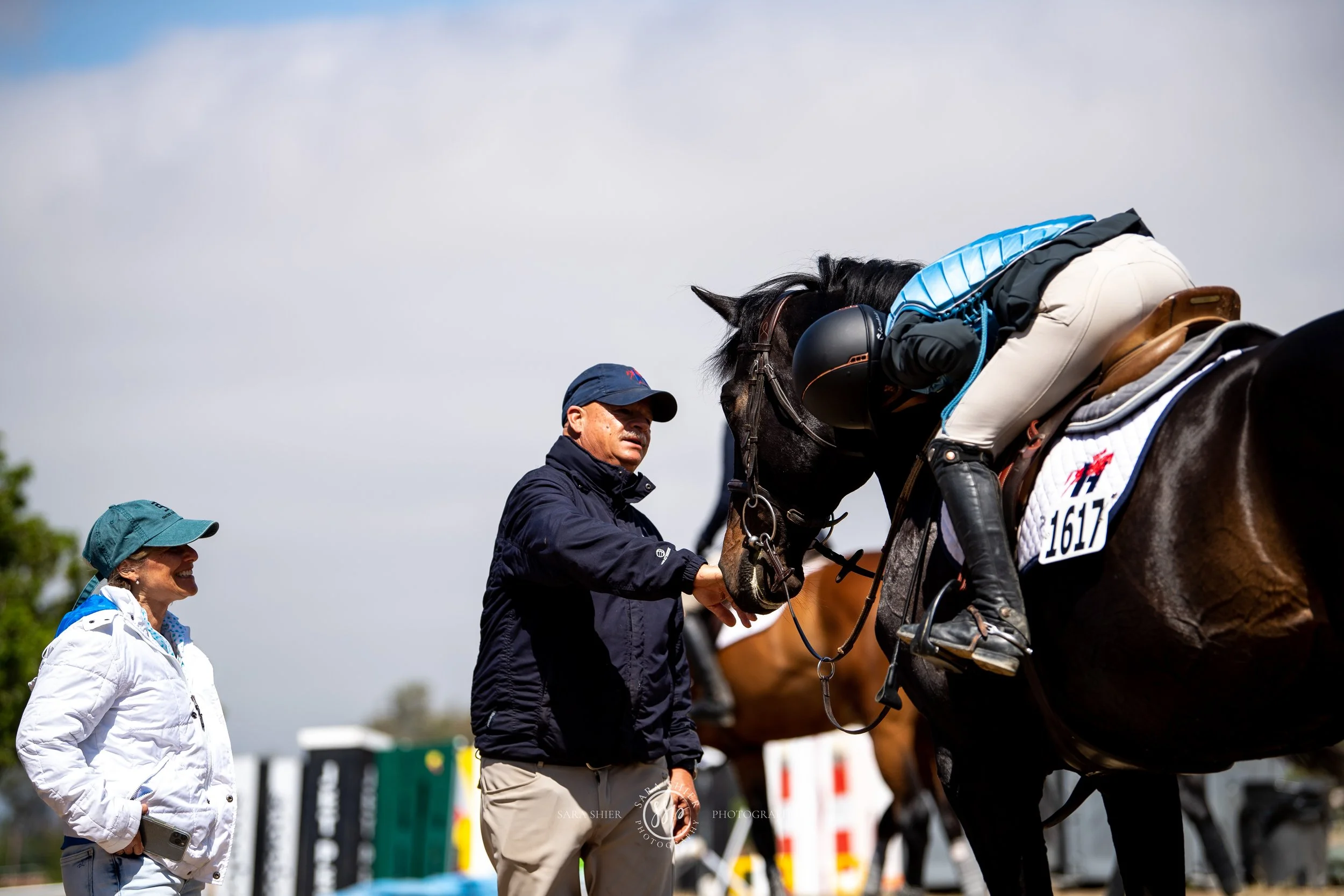 Two women and two men with a horse at a horse riding competition, with the men checking the horse's tack and the women smiling.