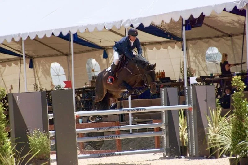 A person riding a horse and jumping over an obstacle at an equestrian event, with a large white tent in the background.