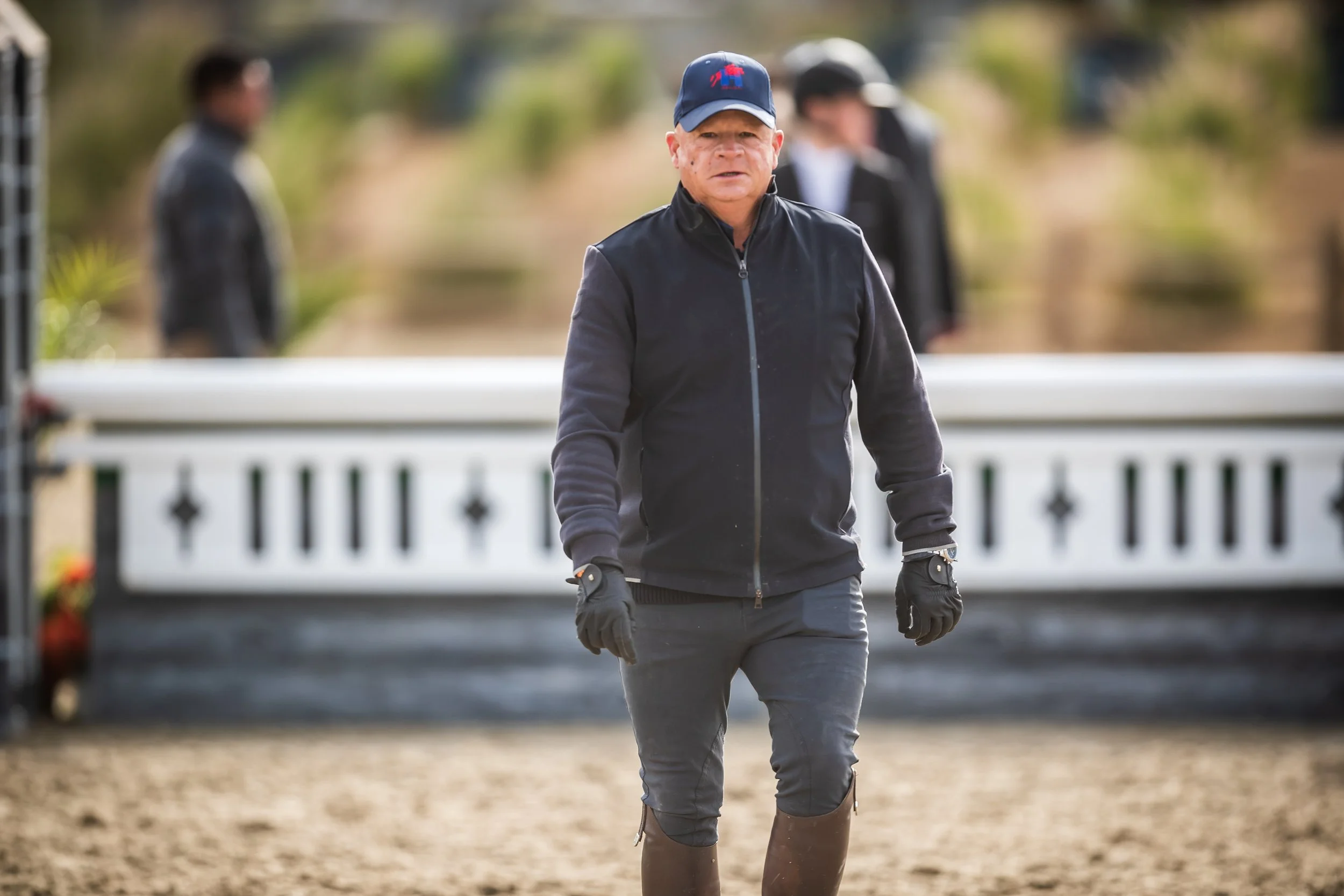 A man dressed in outdoor riding gear, including a cap and gloves, walking on a dirt surface with a white fence and trees in the background.
