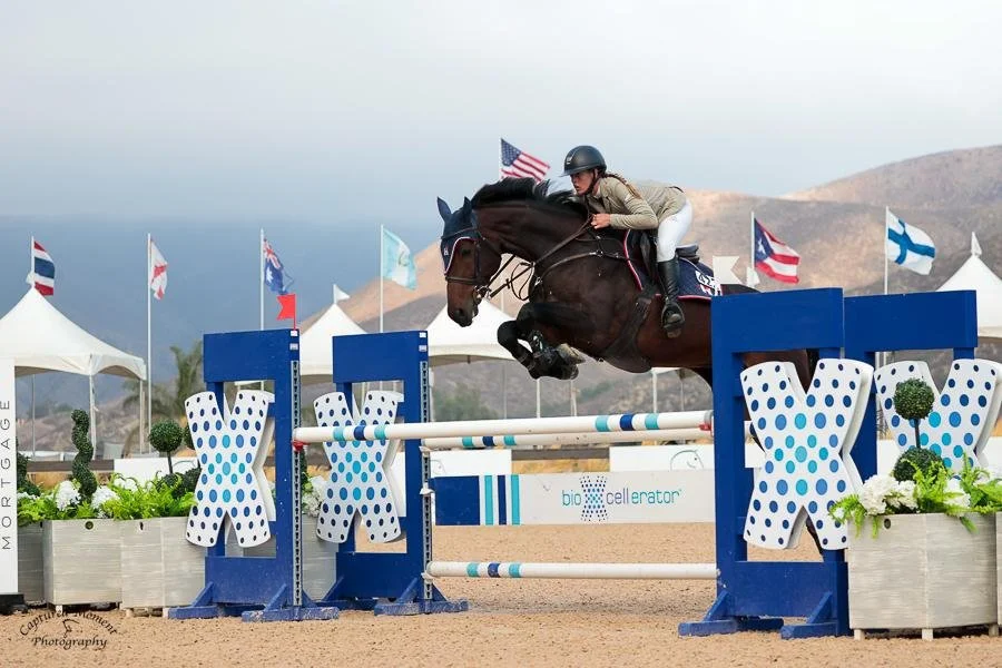 A person riding a horse and jumping over an obstacle in an equestrian event, with tents and flags in the background.