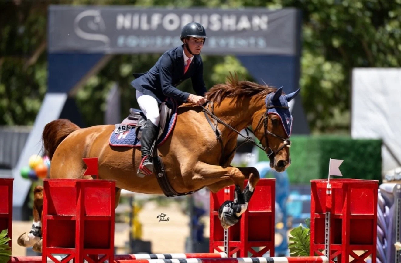 A male equestrian rider in a navy blazer and white breeches guiding a chestnut horse over a red and white jump during a show jumping competition.