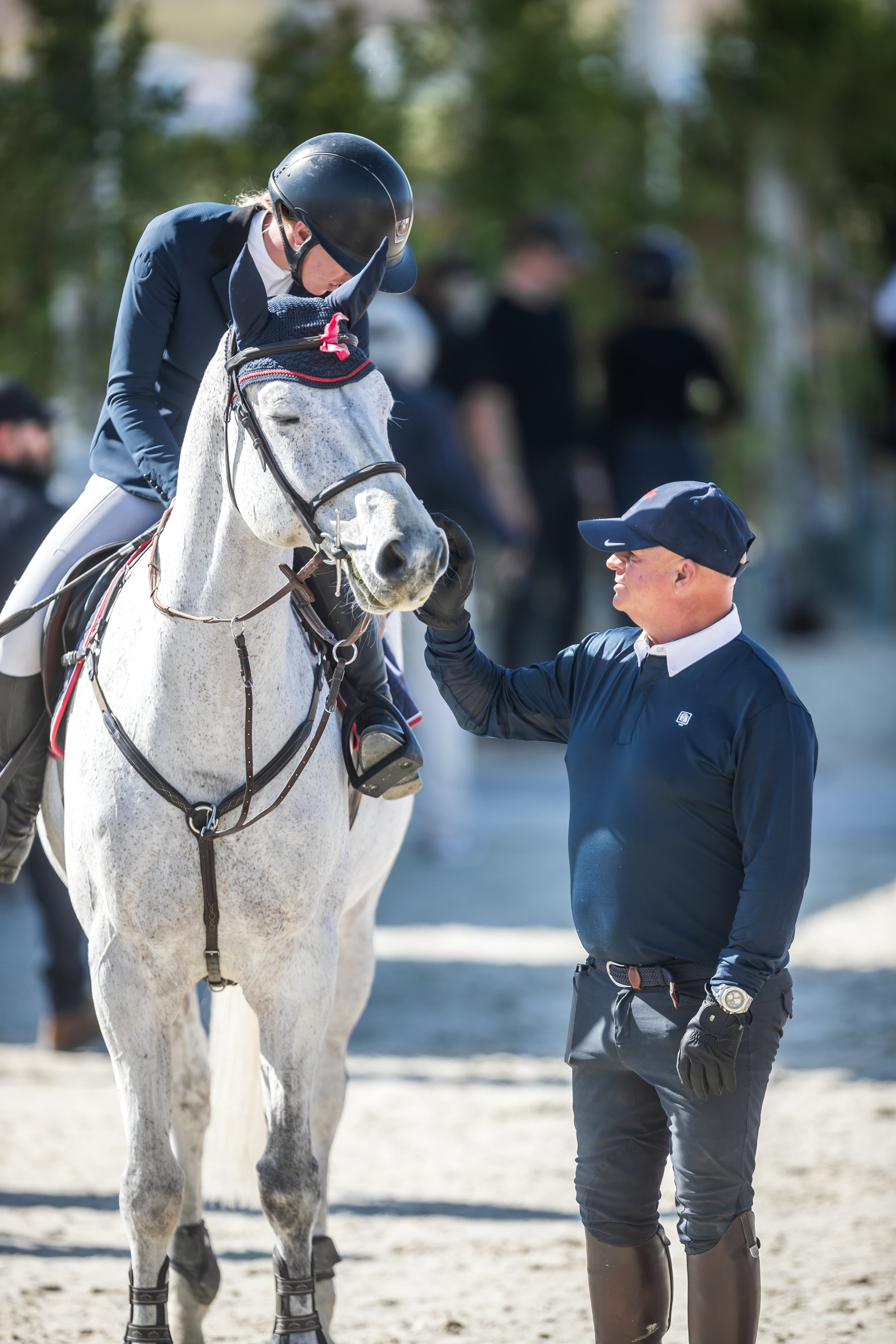 A person in riding gear, wearing a helmet, sitting on a white horse, receives a handshake from another person in a navy blue shirt and a cap, during a daytime equestrian event.
