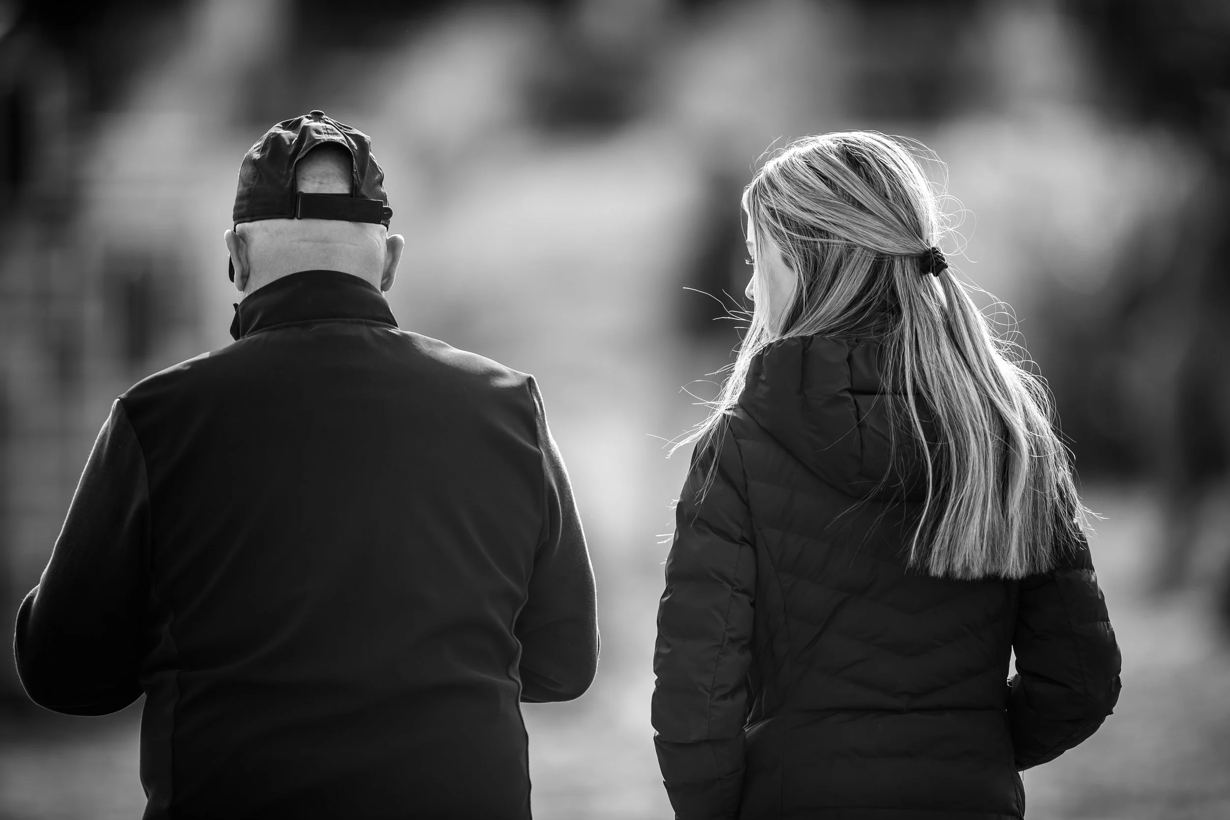 Black and white photo of a man and woman walking side by side outdoors, seen from behind. The man is wearing a cap backwards and a jacket, while the woman has long hair tied back and is wearing a quilted jacket.