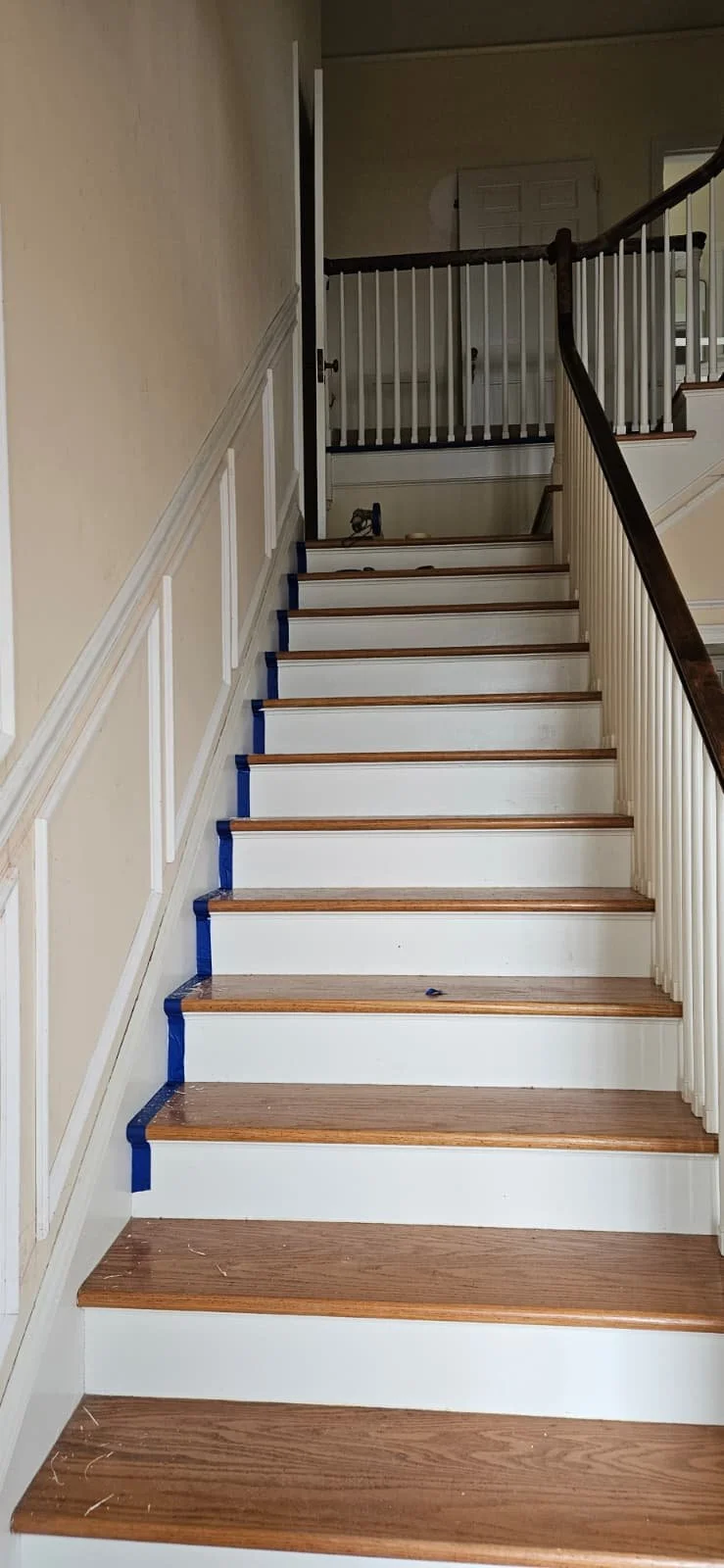 Interior view of a staircase with wooden steps and white risers, partially taped with blue painter's tape, leading up to a landing with a wooden handrail on the right side and a banister with white spindles.