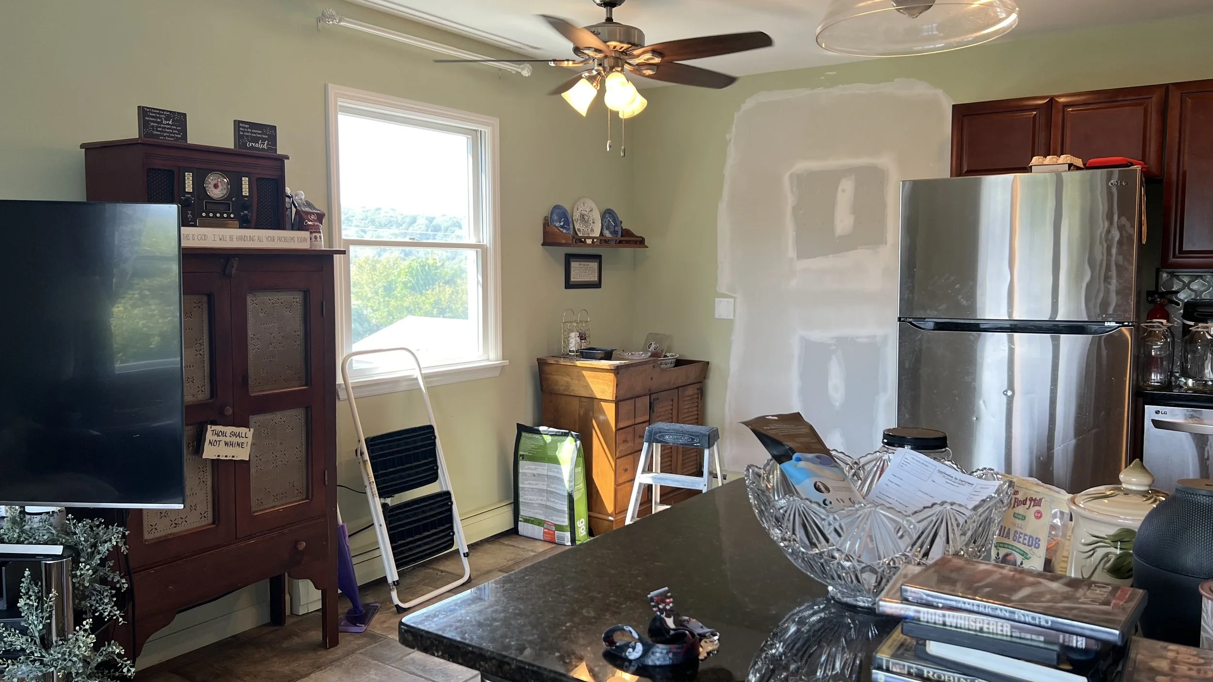 A kitchen with green walls, a window, a ceiling fan with lights, a wooden cabinet, a stainless steel refrigerator, various chairs, a table with miscellaneous items, and some partially painted wall patches.