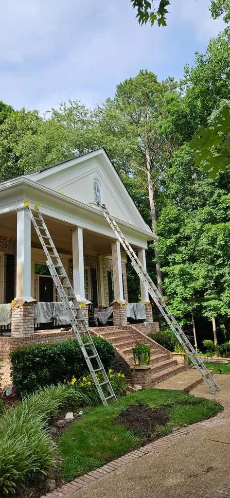 A house under construction with three ladders leaning against the porch, surrounded by green trees and a partly cloudy sky.