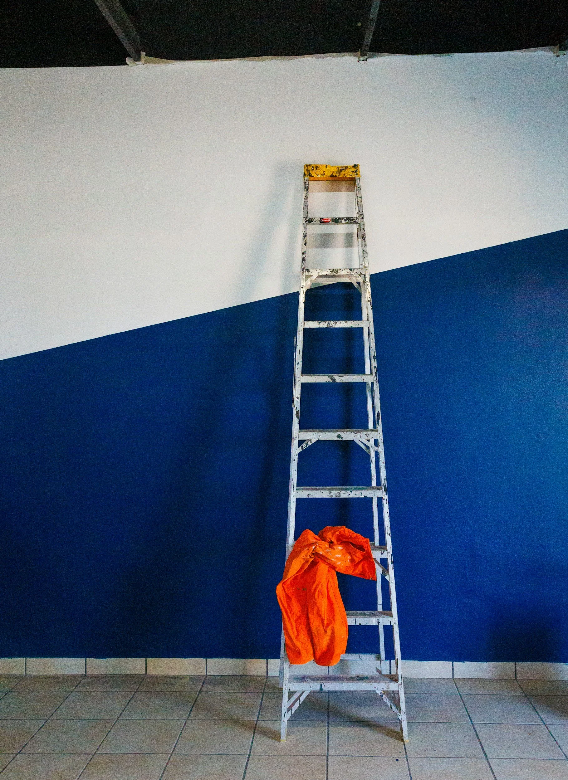 A metal ladder leaning against a wall with a blue lower half and white upper half, with an orange jacket hanging from the bottom rung of the ladder, and a barely visible black ceiling above.