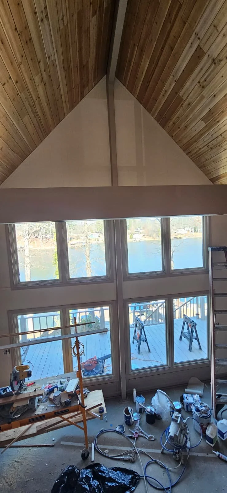 Interior view of a room under construction with a high vaulted wooden ceiling, large windows overlooking water, and construction tools and supplies scattered on the unfinished floor.