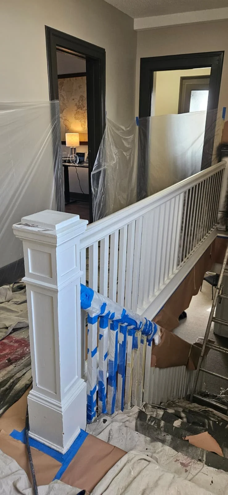 Interior of a house staircase under renovation with white painted balusters, plastic sheeting covering walls, and painter's tape on newel posts and railing, near doorways.