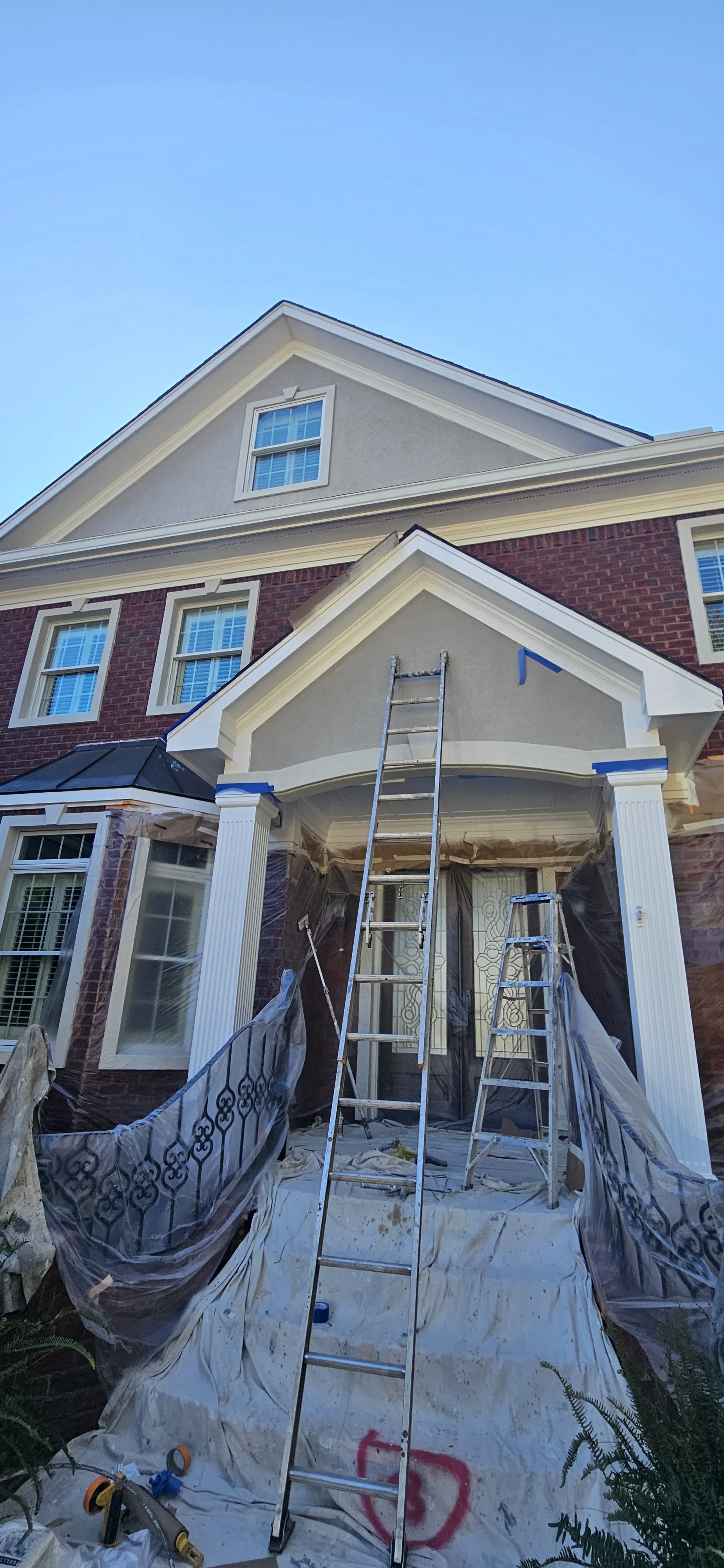 Construction workers are renovating the front porch of a house, with ladders, tools, and plastic sheeting covering parts of the area.