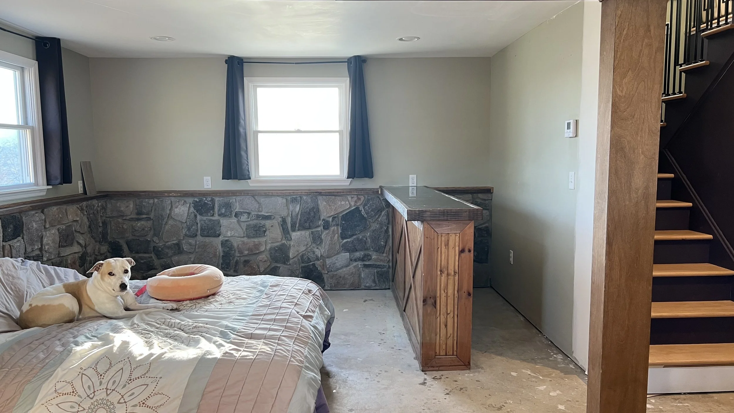 Living room with light green walls, a stone half-wall, and a window with dark blue curtains. A white and tan dog lies on a bed, which has a peach-colored dog bed. There is a small wooden bar or counter, a staircase with wooden steps and black railing