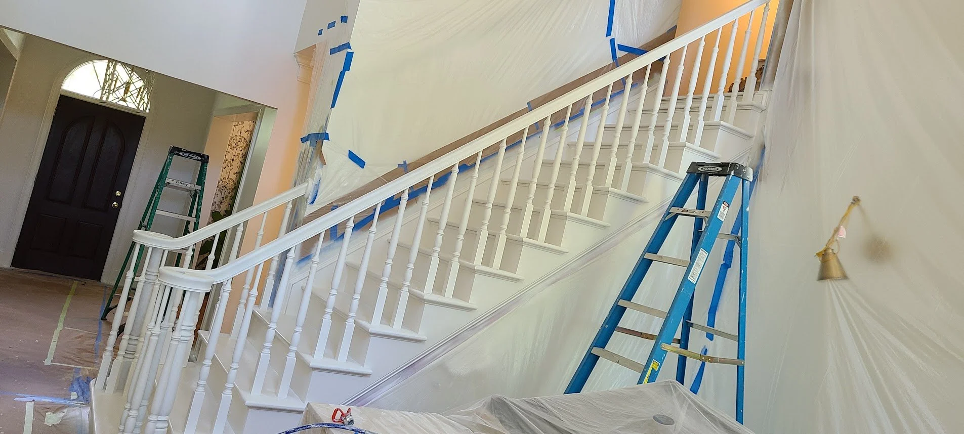 Interior of a house under renovation with a staircase, painter's plastic sheeting covering the floor, blue painter's tape on the walls and trim, and a blue ladder positioned near the staircase.
