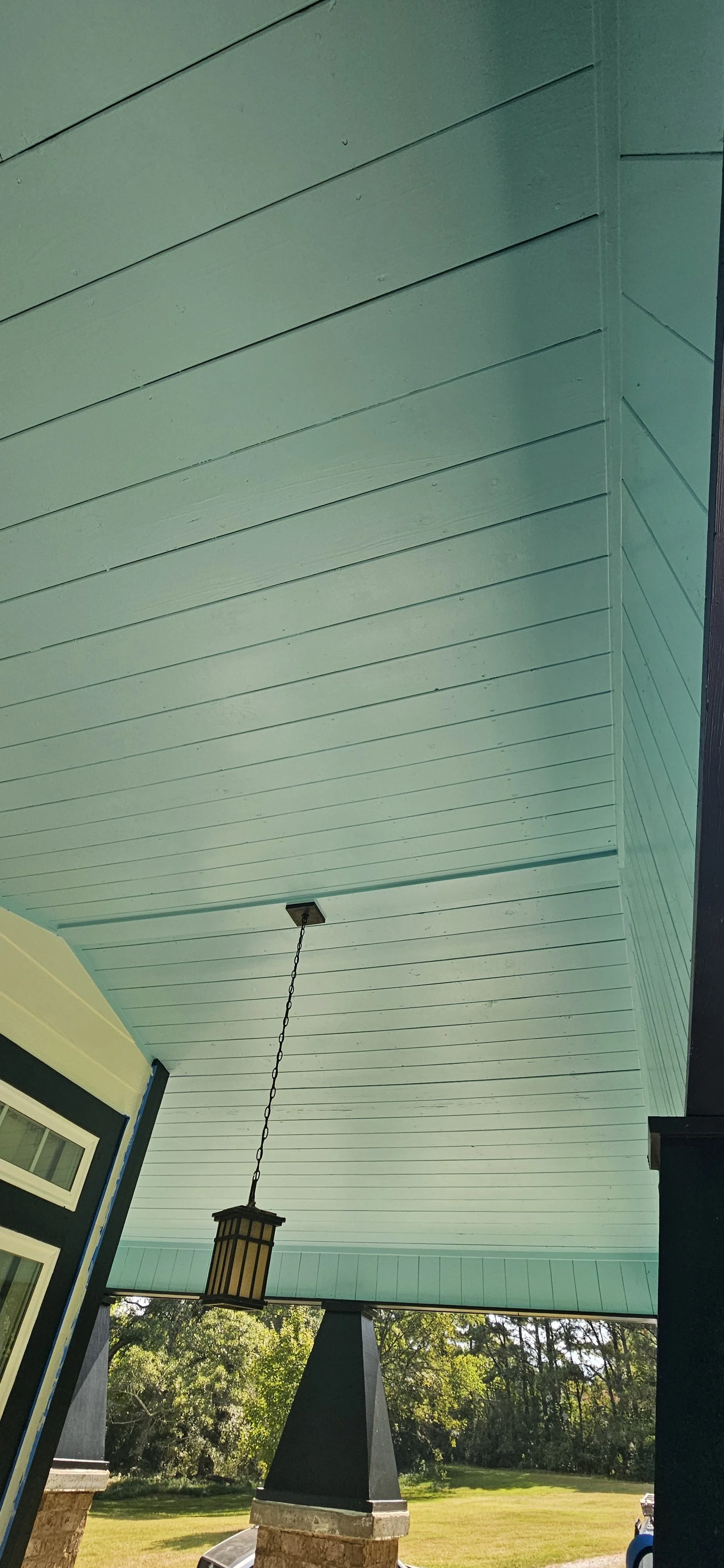 View of a porch ceiling painted light blue, with a hanging lantern-style light fixture and a view of a grassy yard and trees in the background.