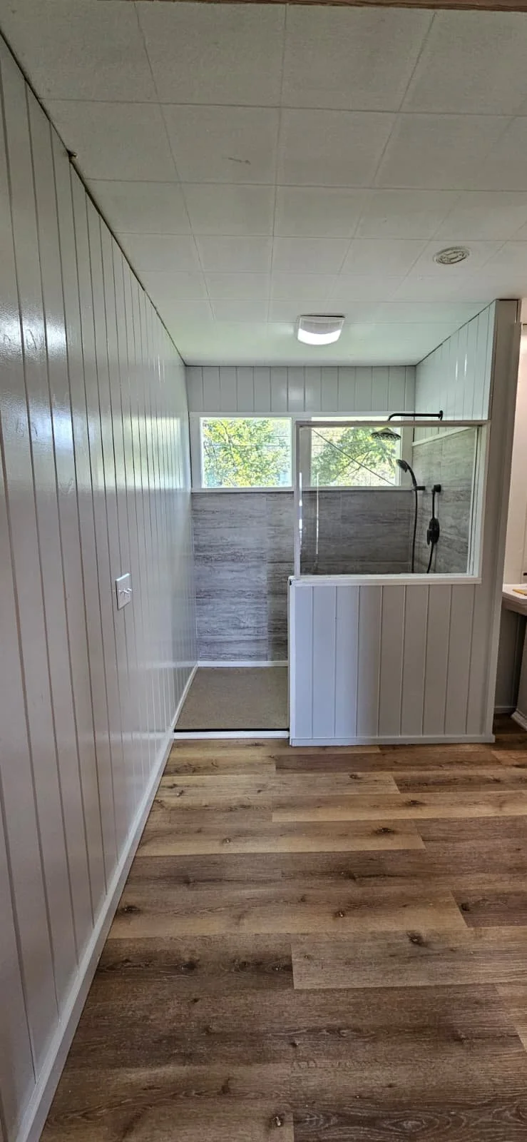 Bathroom with wooden floor, white paneled walls, ceiling light, and a walk-in shower with a glass partition and a window showing trees outside.