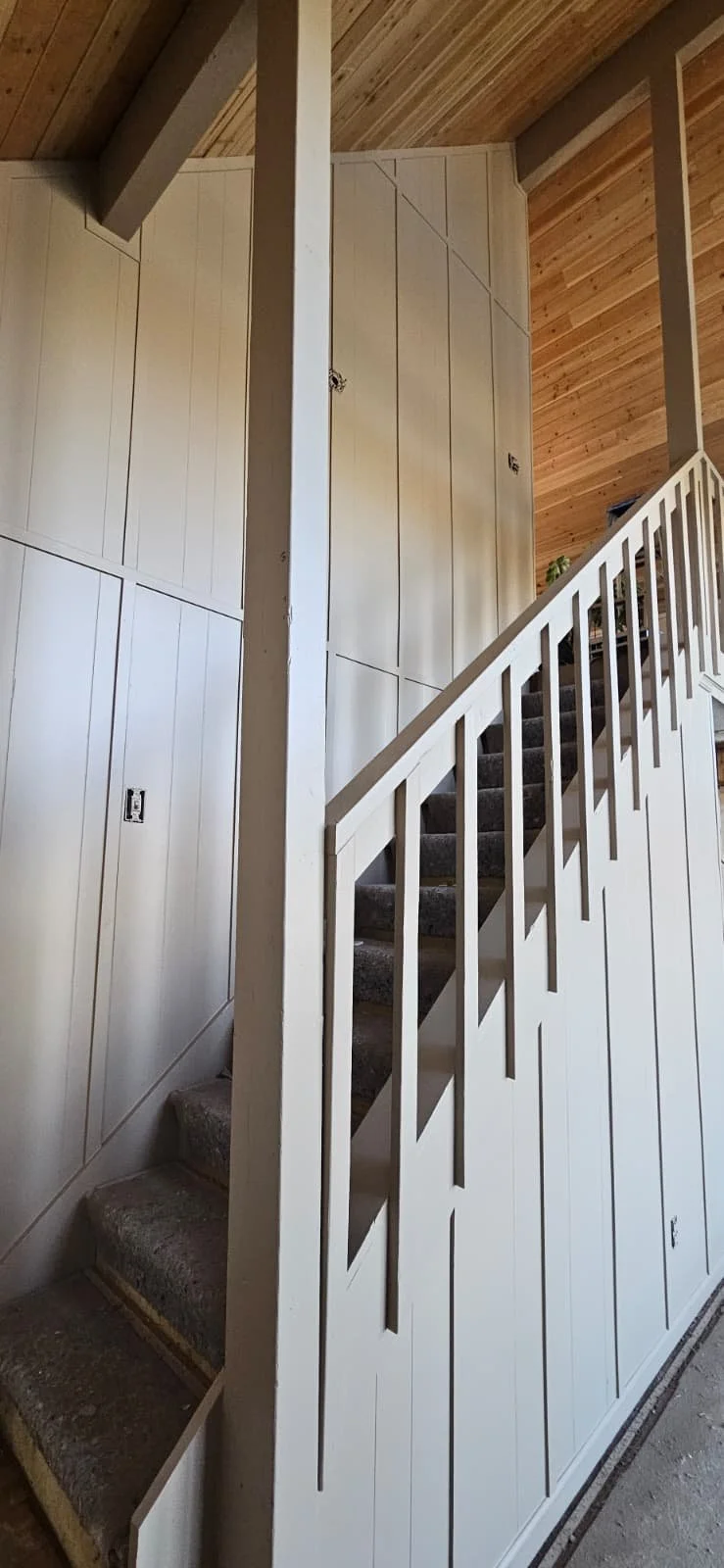 Interior of a house staircase with white paneling, brown carpeted stairs, and wooden ceiling.