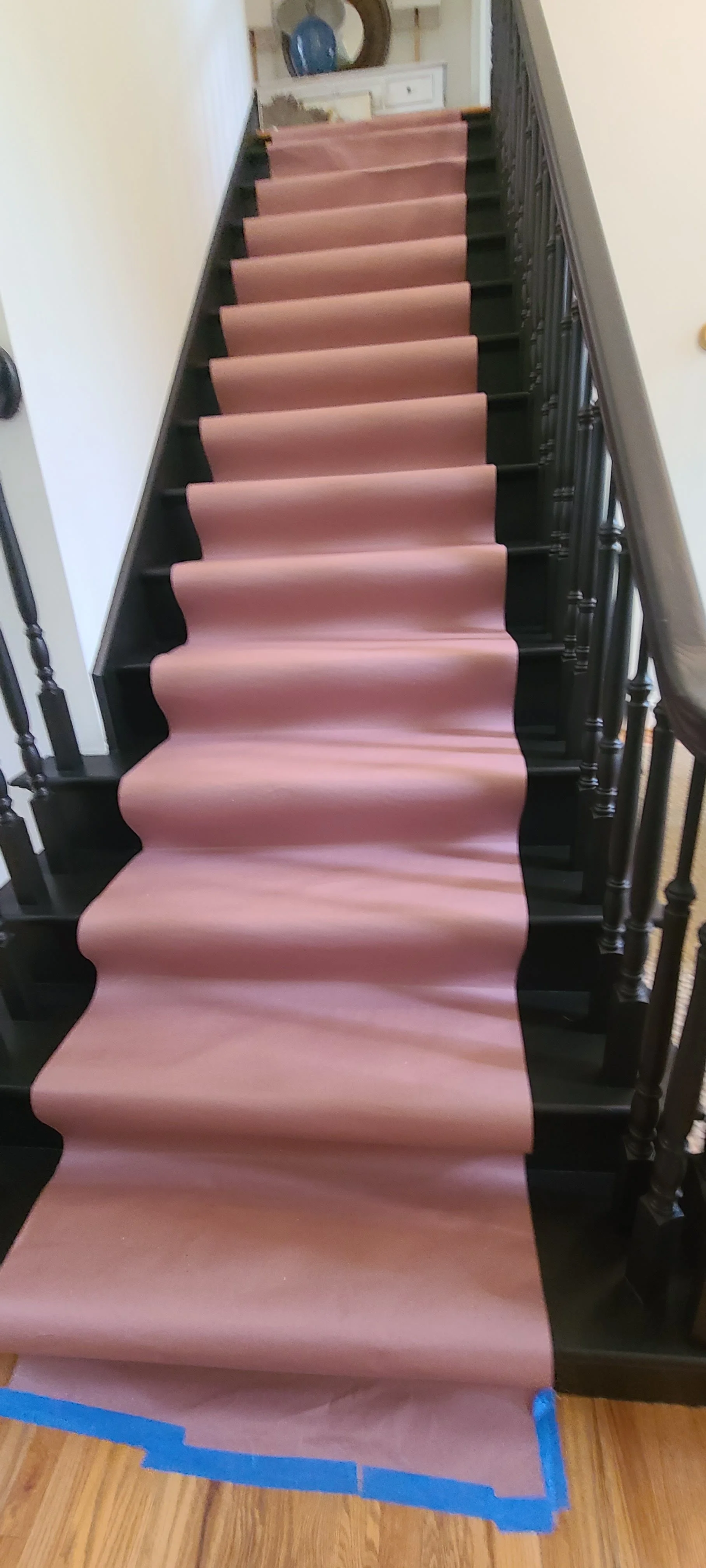 Staircase with black wooden railings and light pink carpet runner, partially covered with blue painter's tape at the bottom.