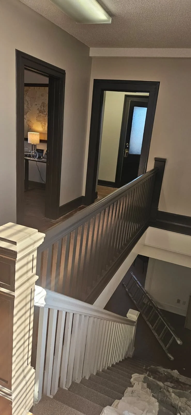 Interior view of a staircase landing with two adjacent dark-framed doorways, one leading to a room with a lamp and map on the wall, and the other to an unknown space, with a wooden staircase and ladder below.