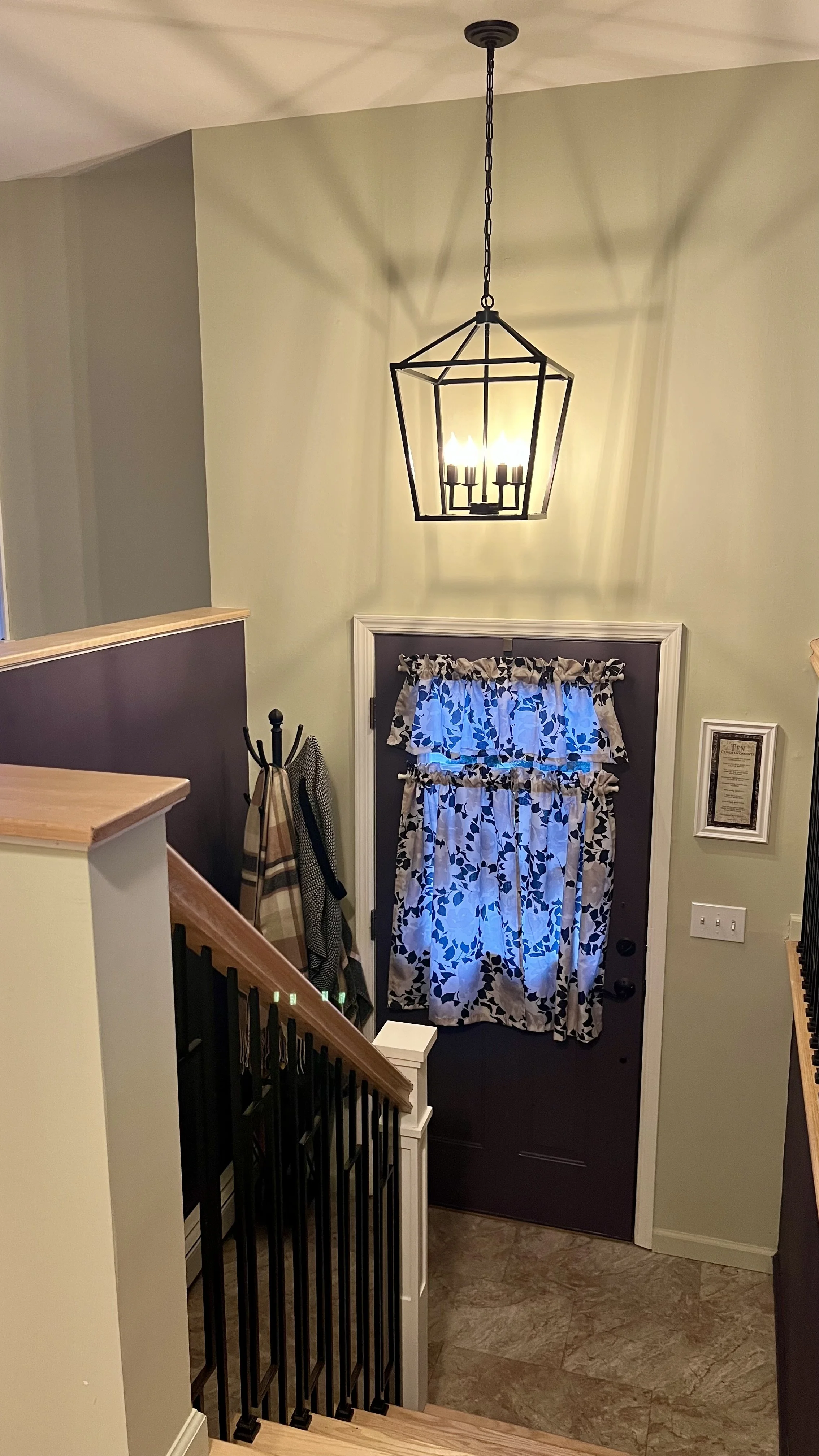 Interior view of a house entryway with a chandelier hanging from the ceiling, a dark-colored door covered with a decorative curtain, and a coat rack with hats and scarves hanging. The stairs and railing are visible in the foreground.