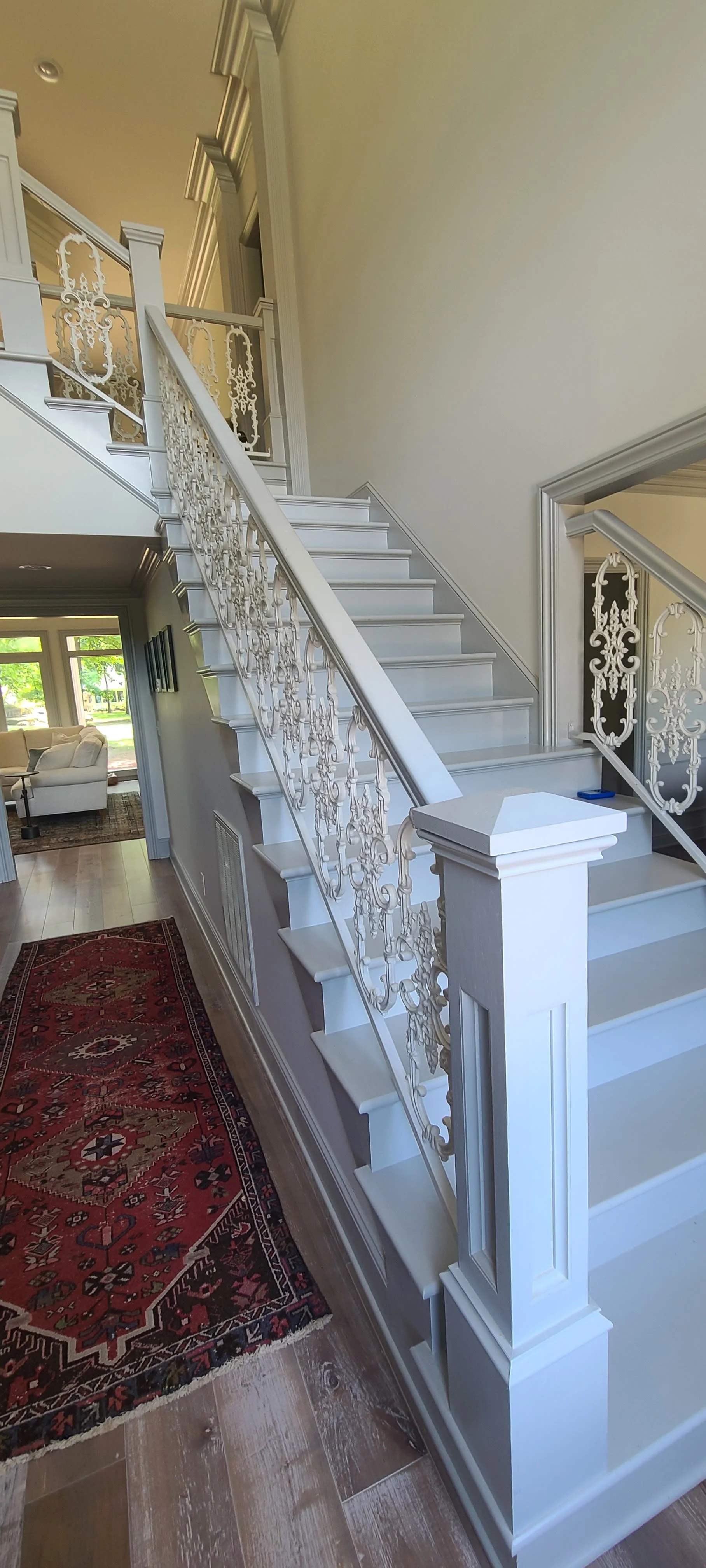 Interior view of a white staircase with decorative iron railings, leading to an upper floor, in a well-lit home with hardwood floors and a red patterned area rug.