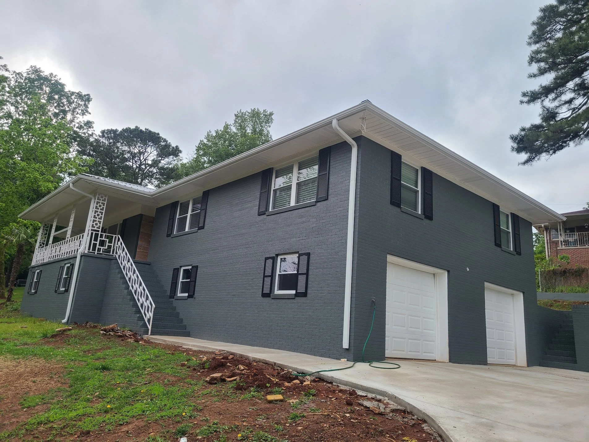 A two-story gray house with black window shutters and a white garage door. There is a staircase with white railings leading to the front door. The house is surrounded by green trees and an overcast sky.