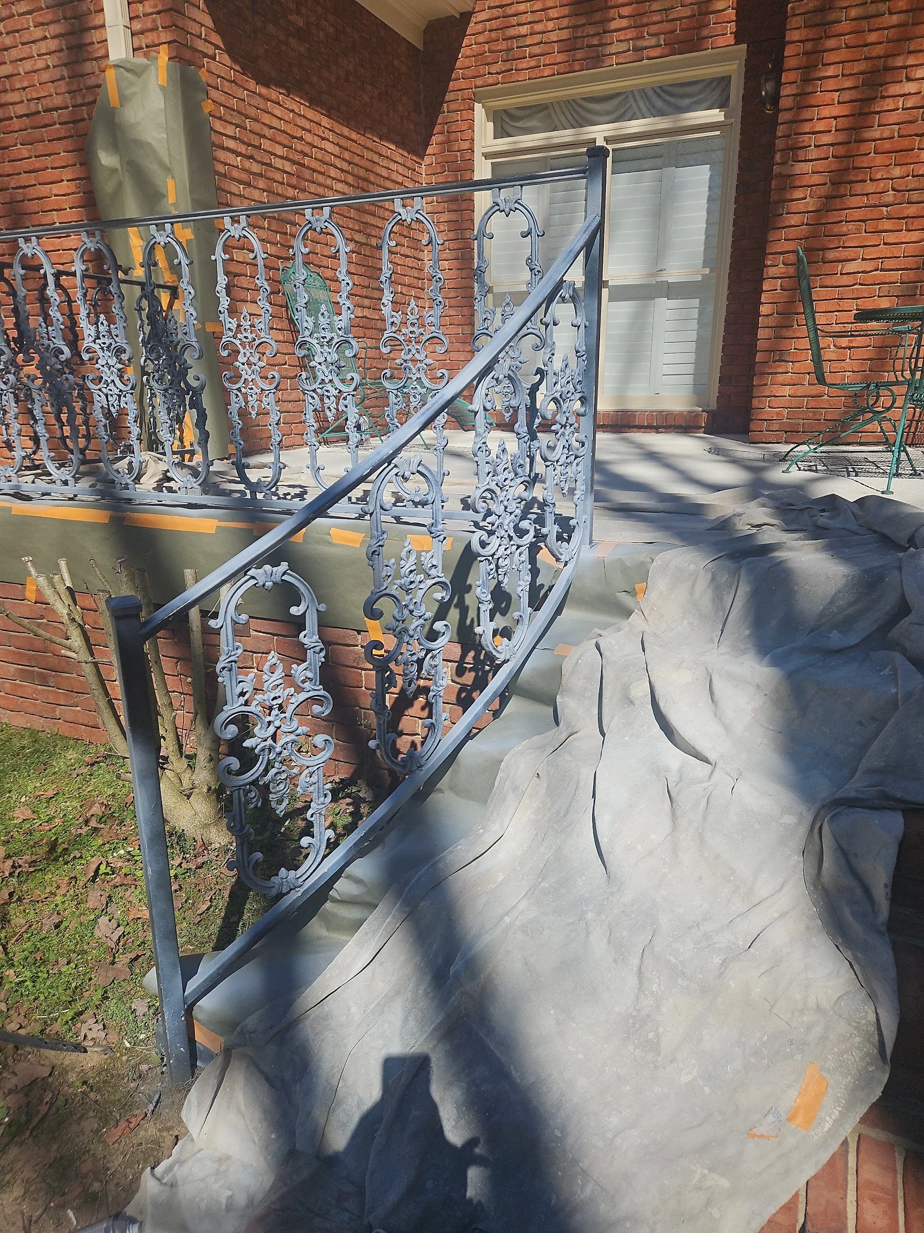 A decorative wrought-iron railing on a brick building's porch or balcony, with protective covering on the floor and some fall leaves around.