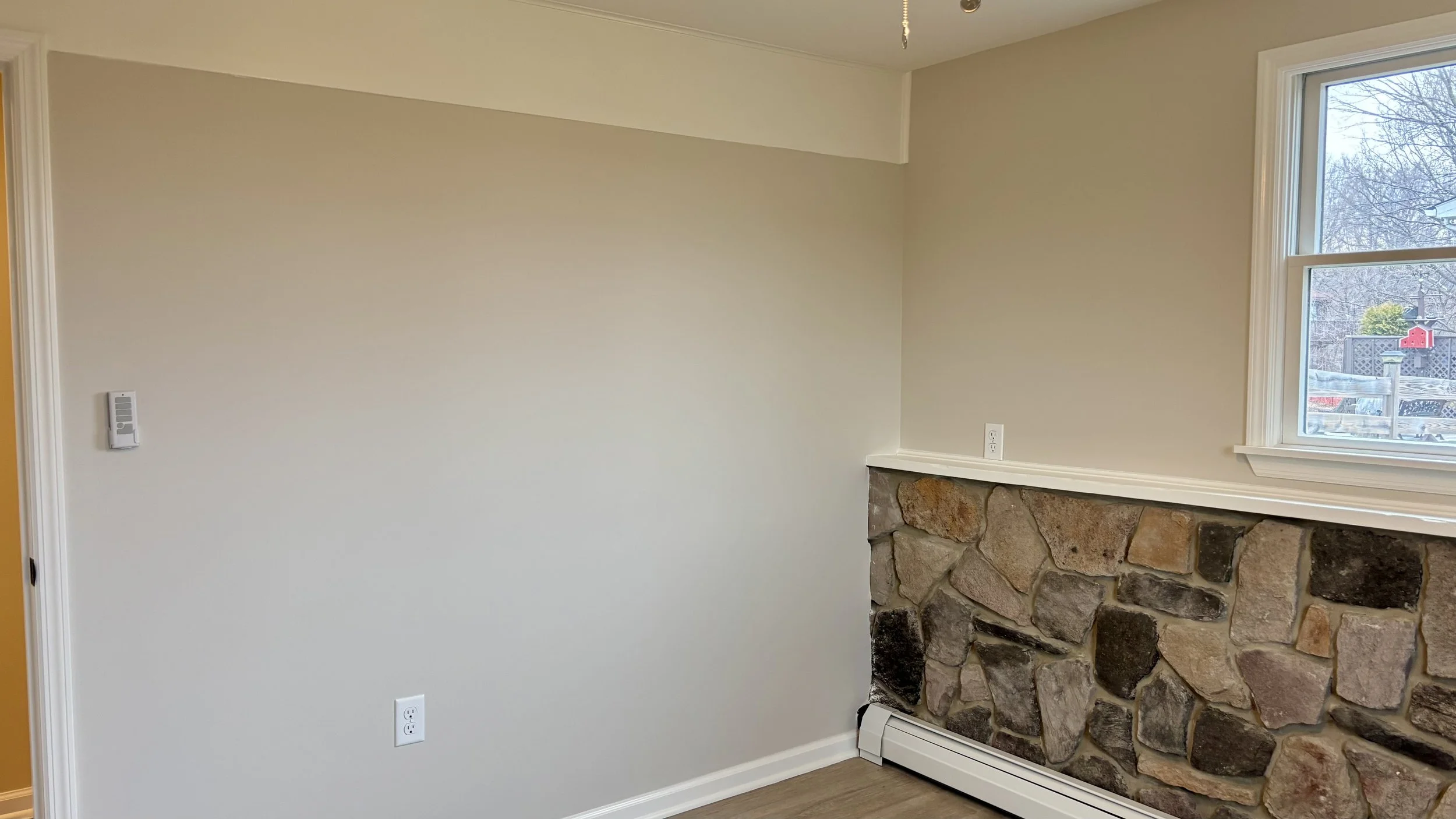 Interior view of a room with a stone fireplace, a large window showing an outdoor scene with trees and a fence, and blank painted walls with electrical outlets.