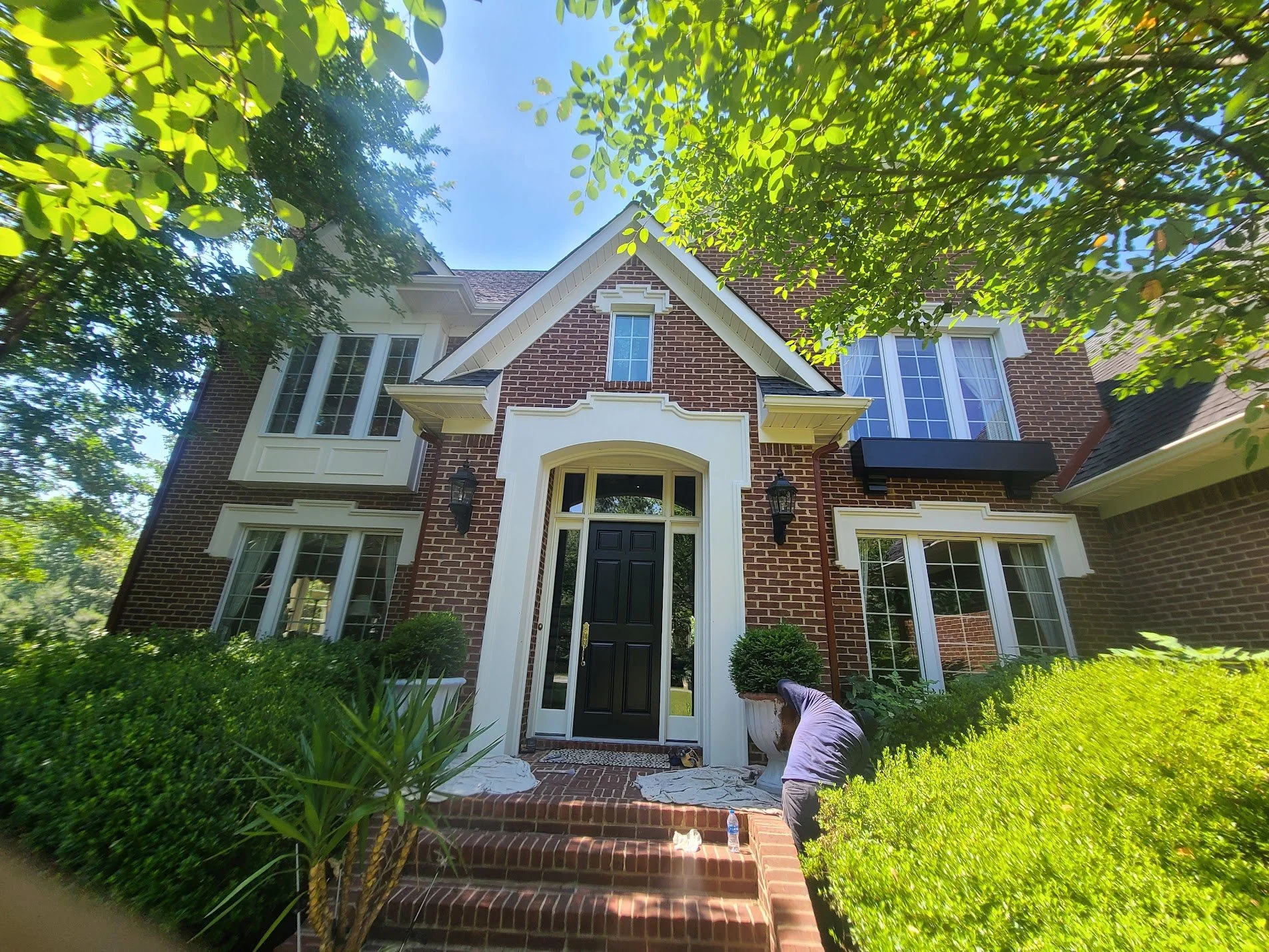A two-story brick house with white trim, a black front door, and multiple windows is surrounded by lush green trees and bushes. A person is working on the steps leading to the front entrance, which are under renovation.