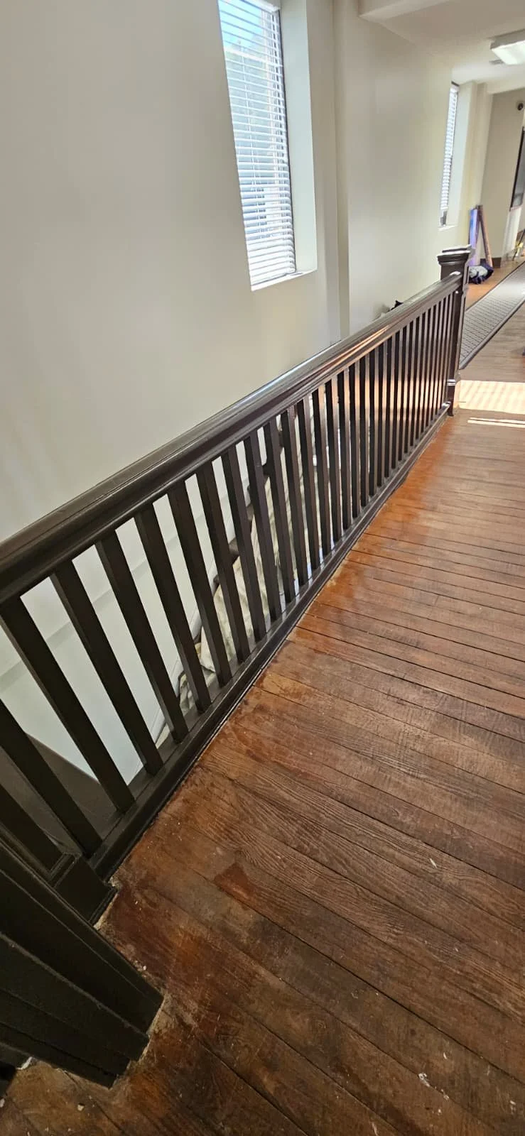 A wooden railing along a staircase, with wooden flooring and windows with blinds on a white wall inside a house.